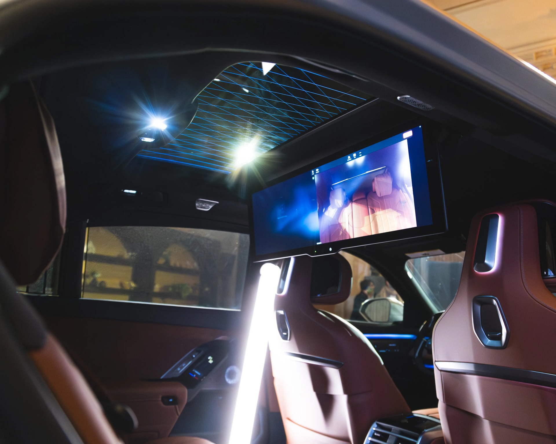 Car interior showing brown leather seats with integrated headrests and a large overhead screen displaying a seat image.