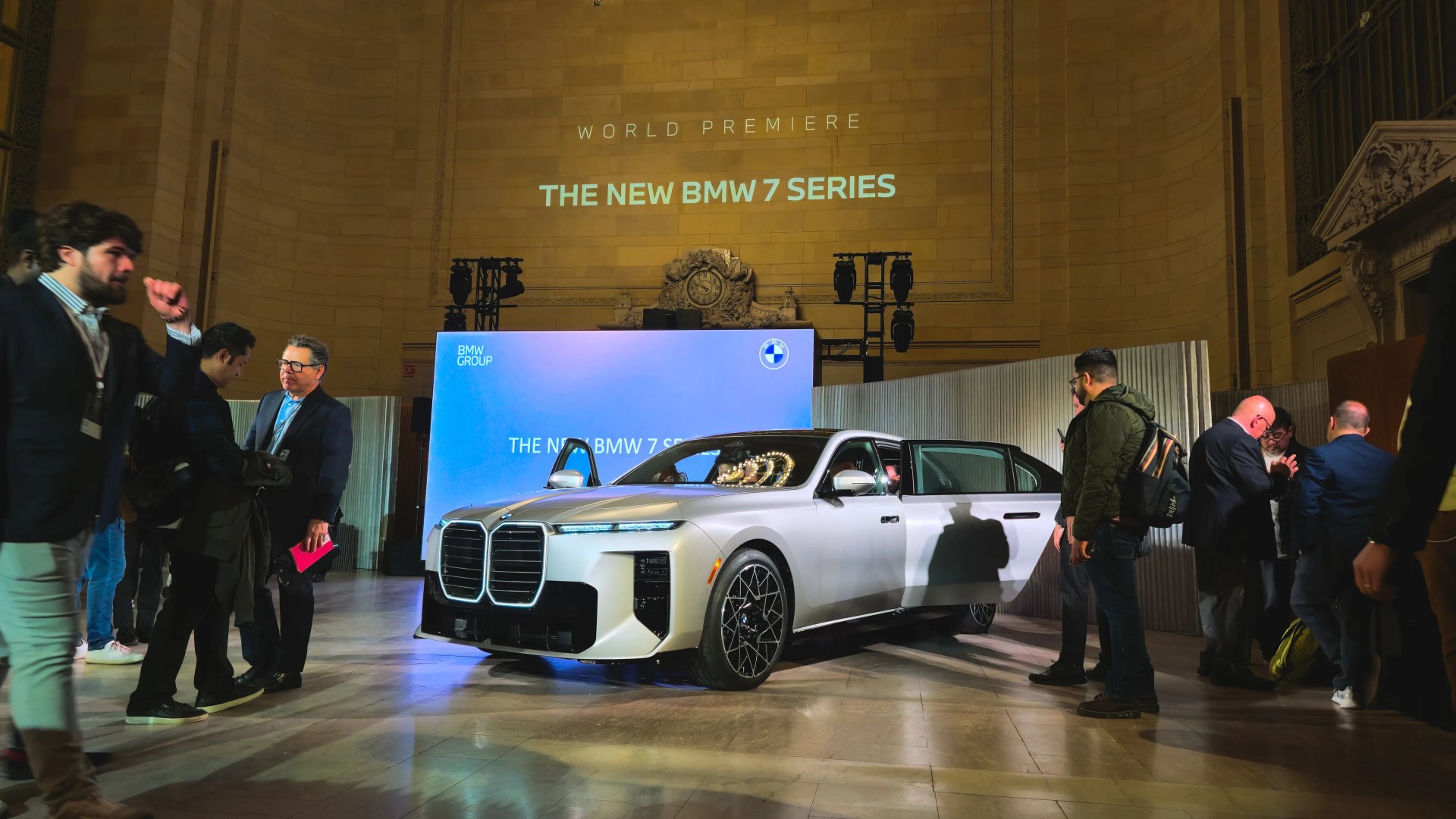Silver BMW 7 Series sedan with open doors displayed indoors at a world premiere event with people around.