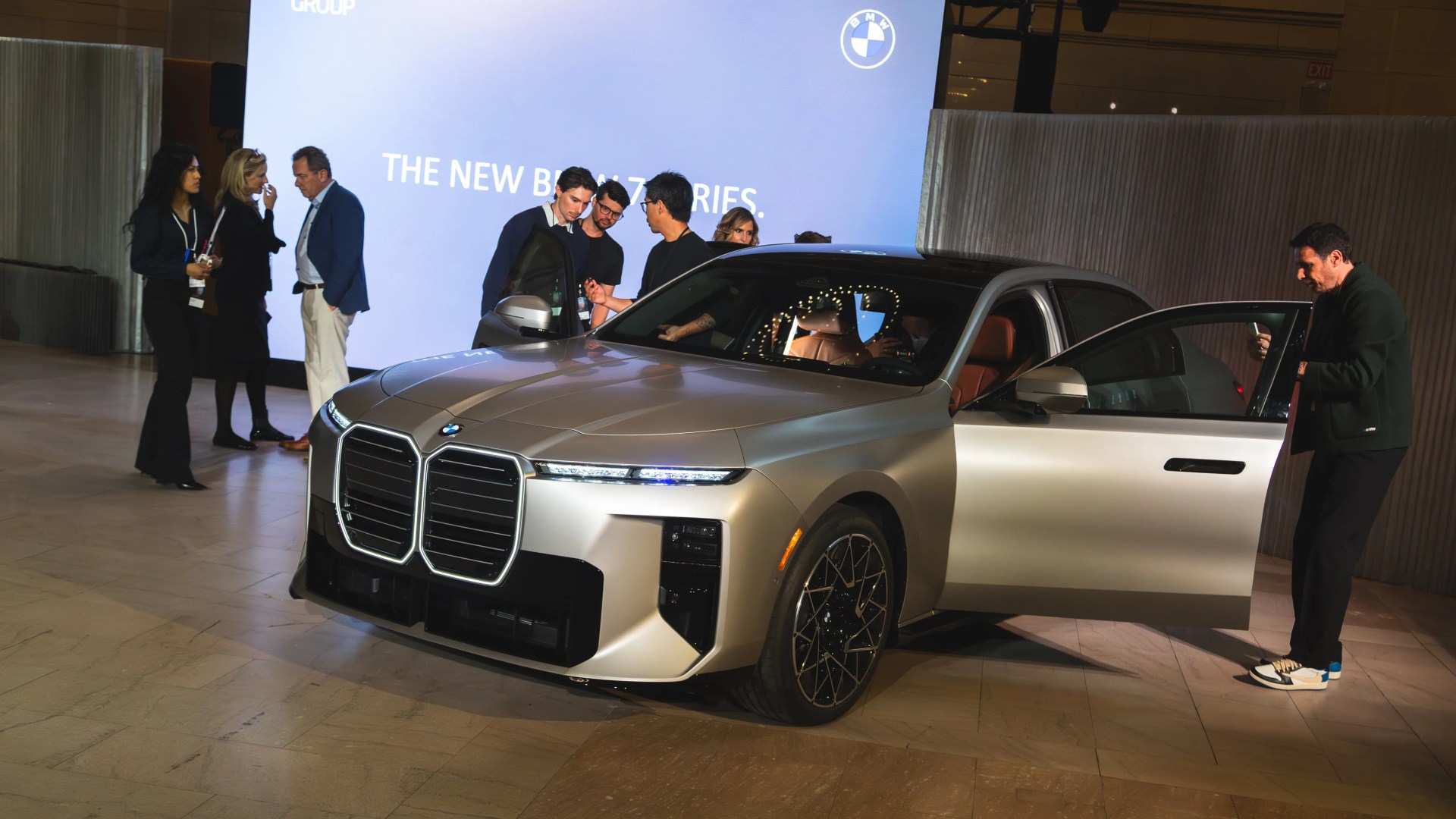 Silver BMW 7 Series sedan with black roof and open doors at an indoor event with people around.