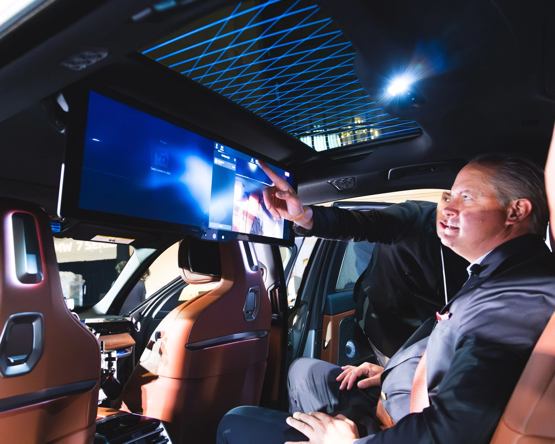 Interior of a luxury car with brown leather seats, a large digital display screen, and a panoramic sunroof with blue grid lighting.