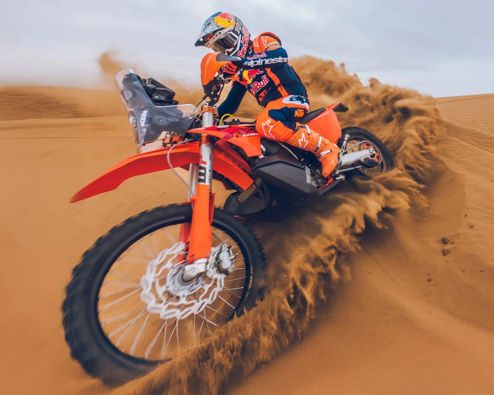Orange KTM dirt bike kicking up sand with rider in matching orange gear and Red Bull helmet in desert.
