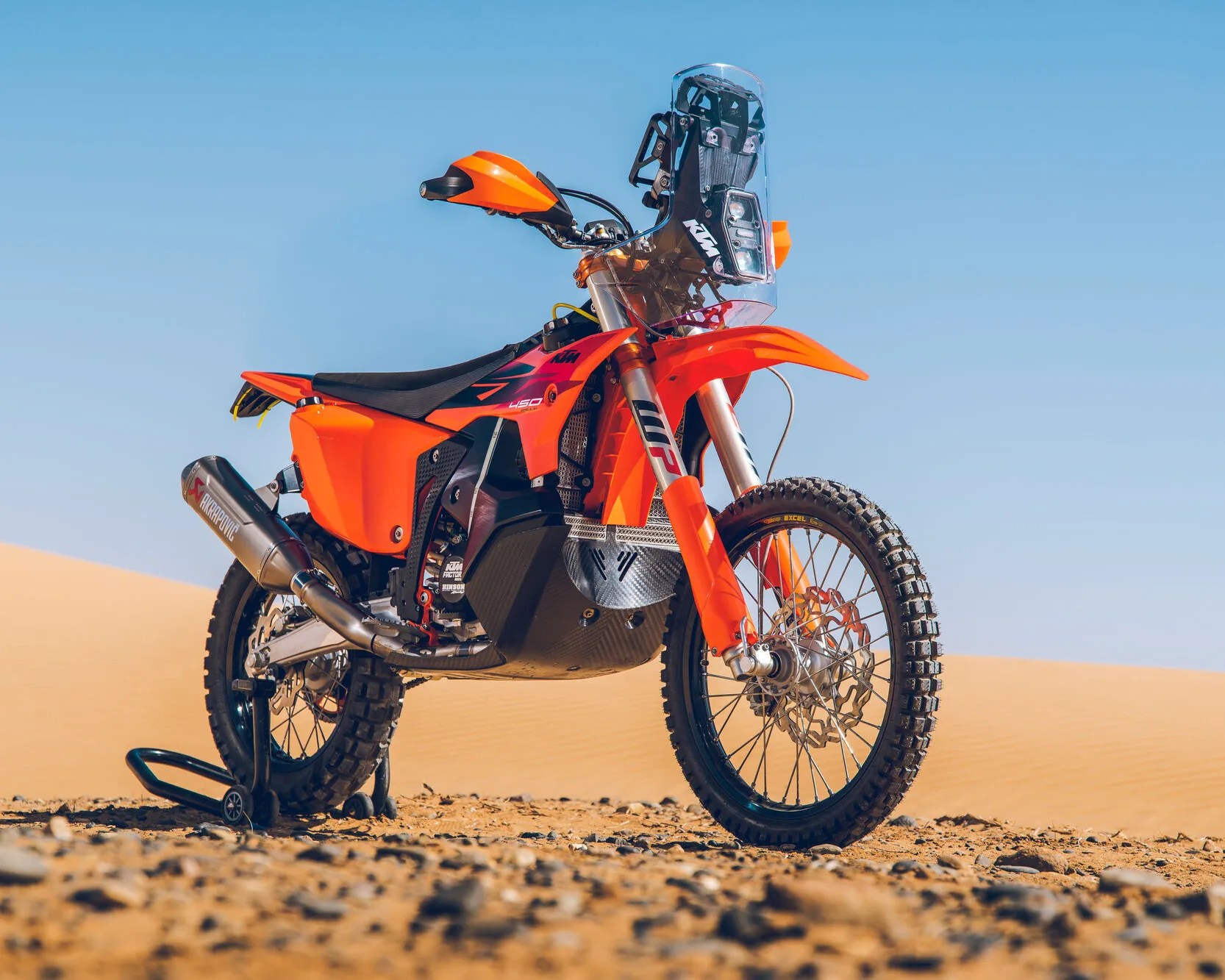 Orange KTM off-road motorcycle with knobby tires and Akrapovic exhaust on rocky desert ground.