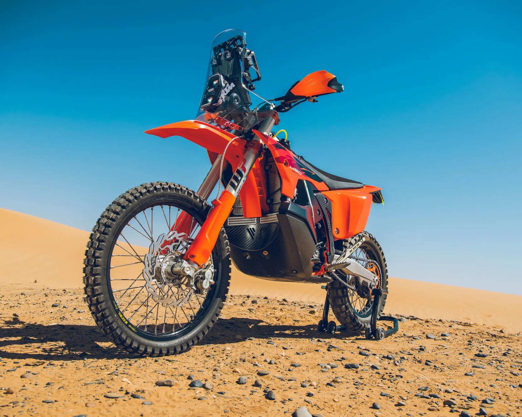 Orange KTM off-road motorcycle with knobby tires on a sandy desert terrain under a clear blue sky.