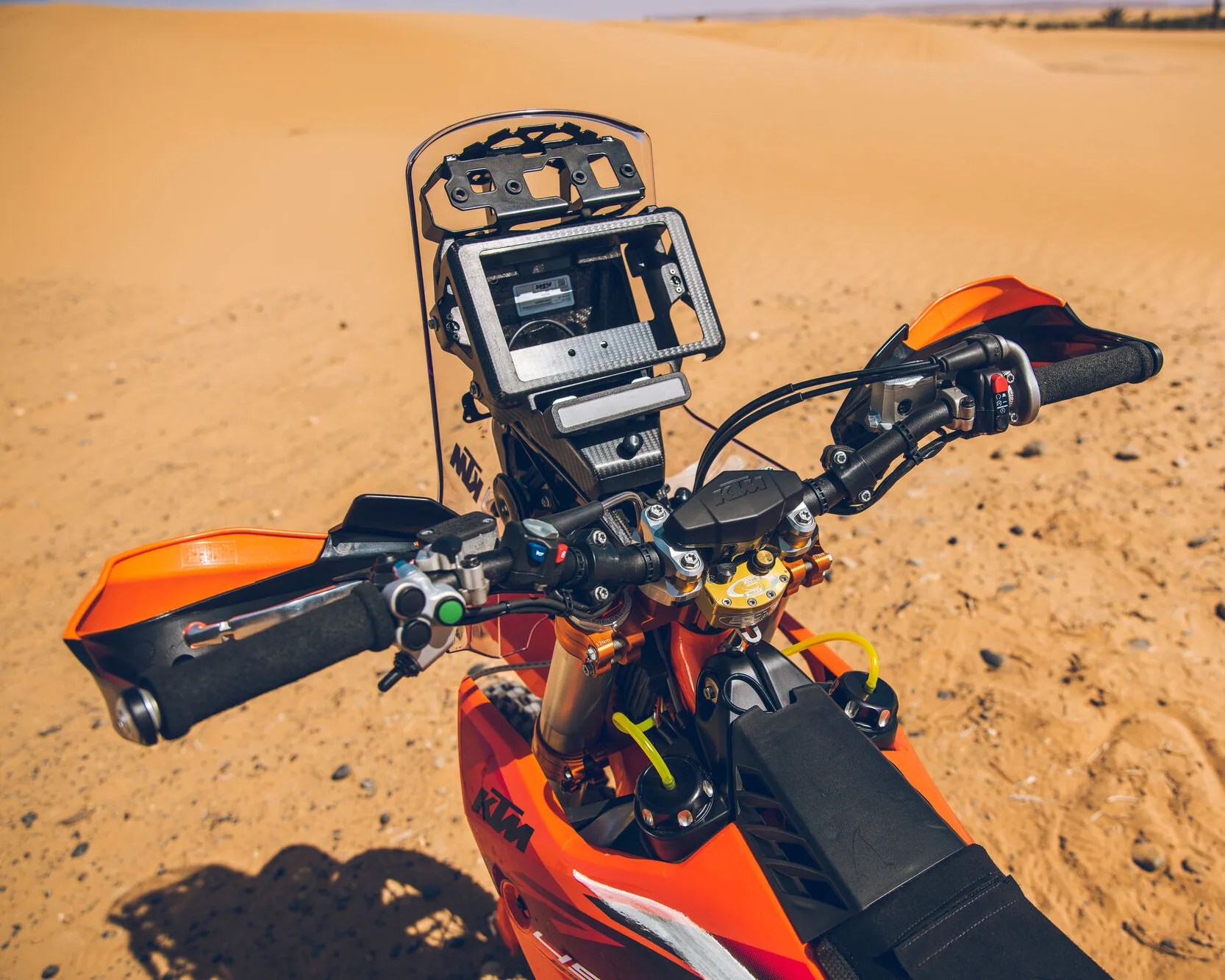 Orange KTM dirt bike handlebar with digital navigation mount and handguards in a sandy desert.