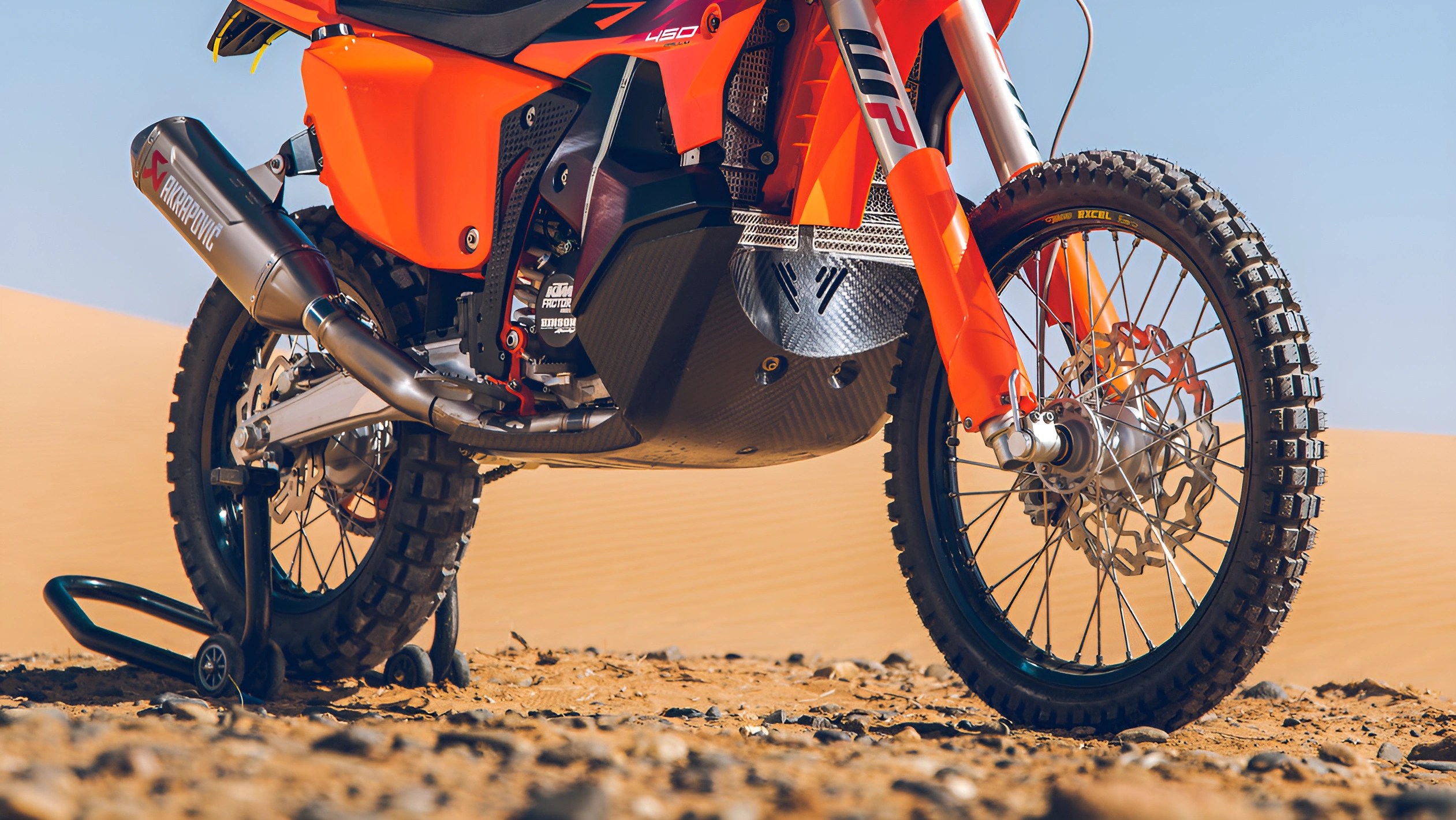 Close-up of an orange off-road motorcycle's front and rear wheels with knobby tires on rocky desert ground.