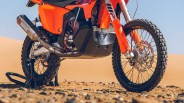 Close-up of an orange off-road motorcycle's front and rear wheels with knobby tires on rocky desert ground.