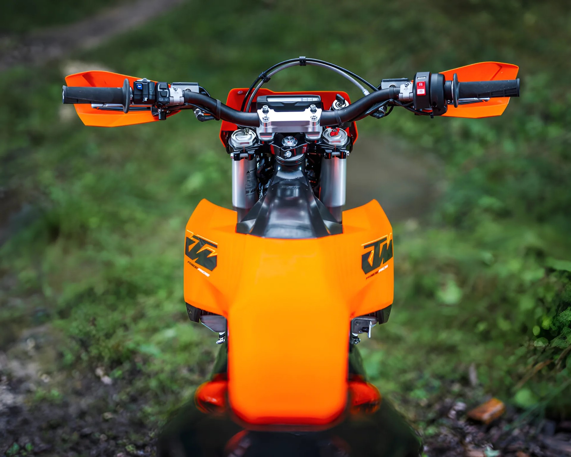 Orange KTM dirt bike handlebars and front panel with controls, viewed from above against a blurred green background.