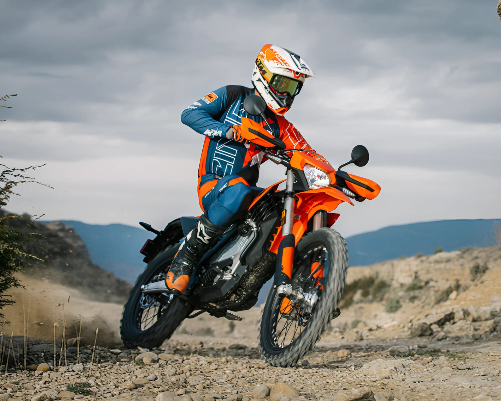 Person wearing blue and orange gear riding an orange off-road motorcycle on a rocky trail.