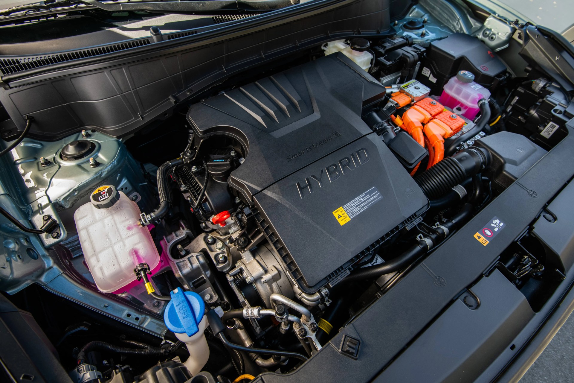 Hybrid car engine compartment with black plastic engine cover labeled "HYBRID" and orange high-voltage cables.