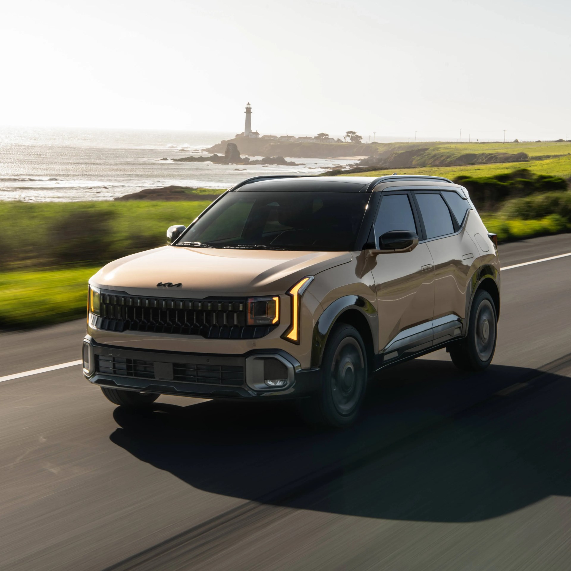 Beige Kia SUV with black roof and illuminated vertical LED headlights driving on a coastal road with a lighthouse in the background.