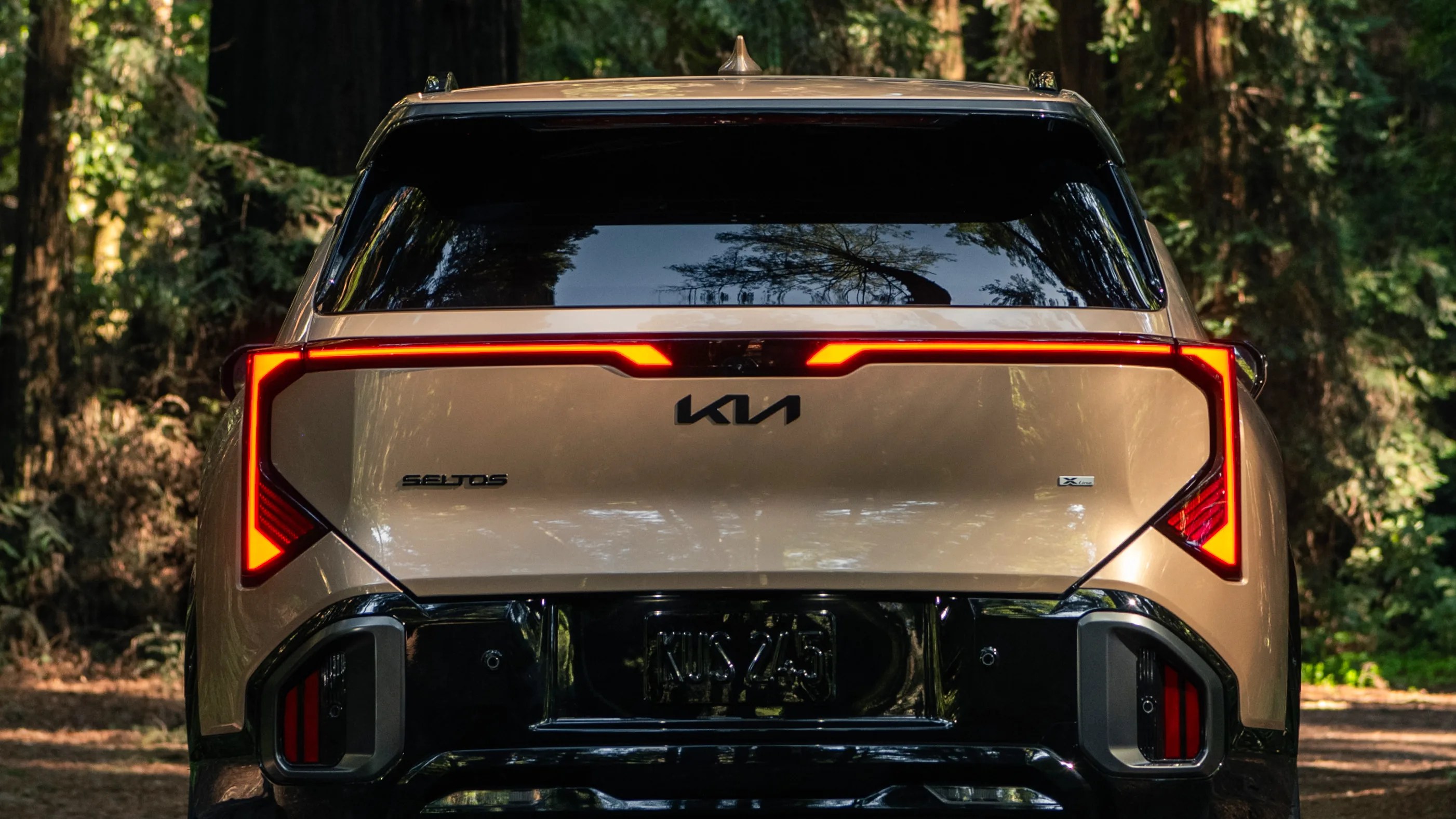 Rear view of a gold Kia Seltos X-Line with illuminated angular taillights in a forest setting.