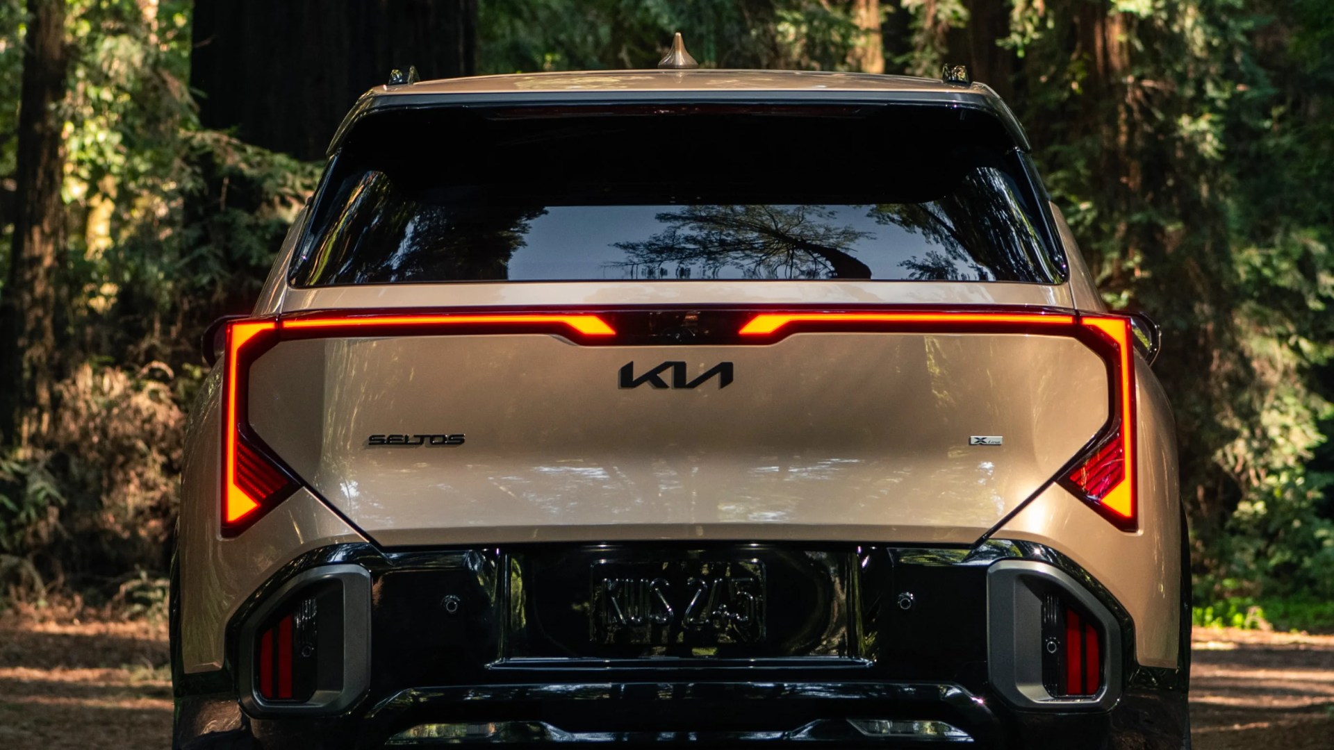 Rear view of a gold Kia Seltos X-Line with illuminated angular taillights in a forest setting.