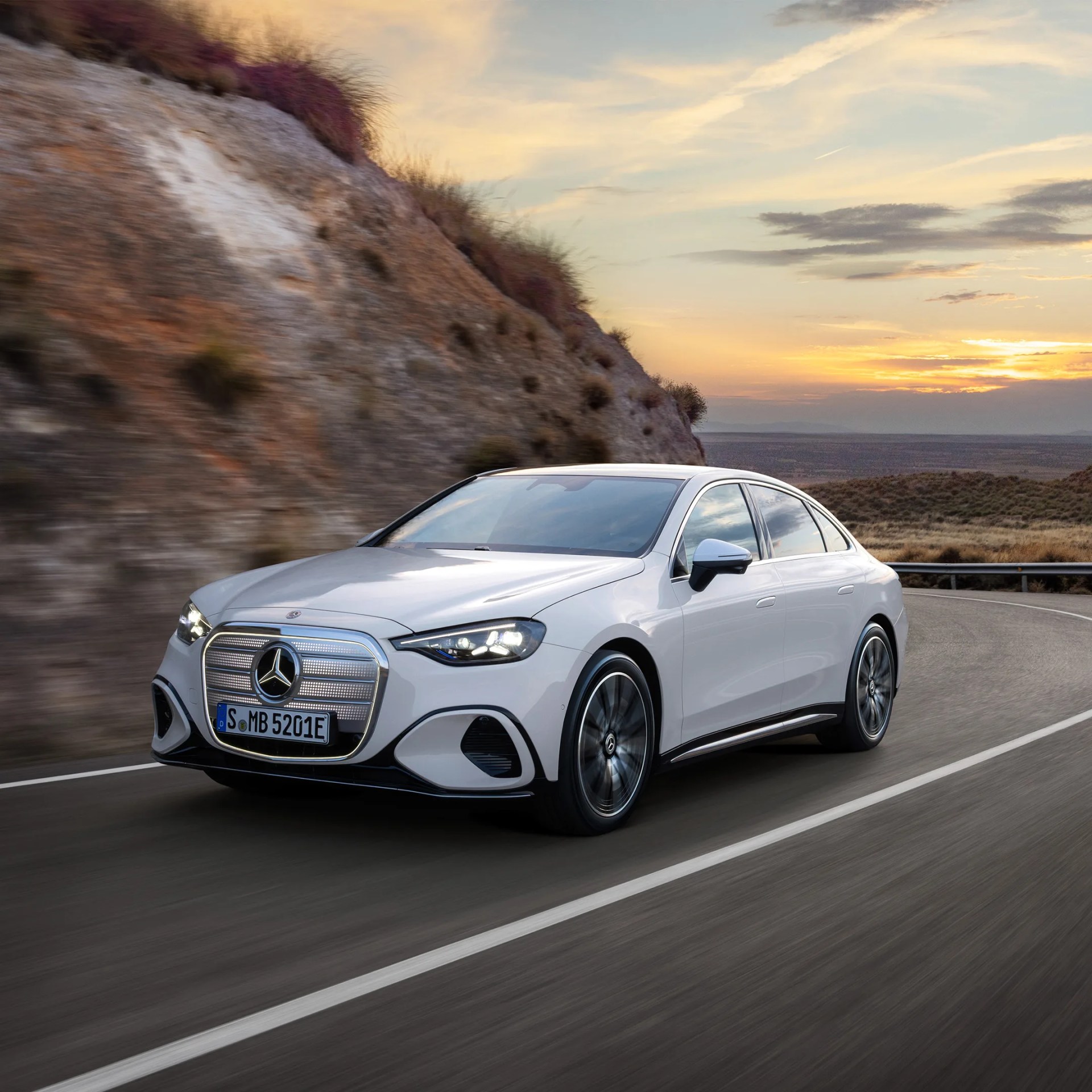 White Mercedes-Benz electric sedan driving on a winding road at sunset with illuminated headlights.