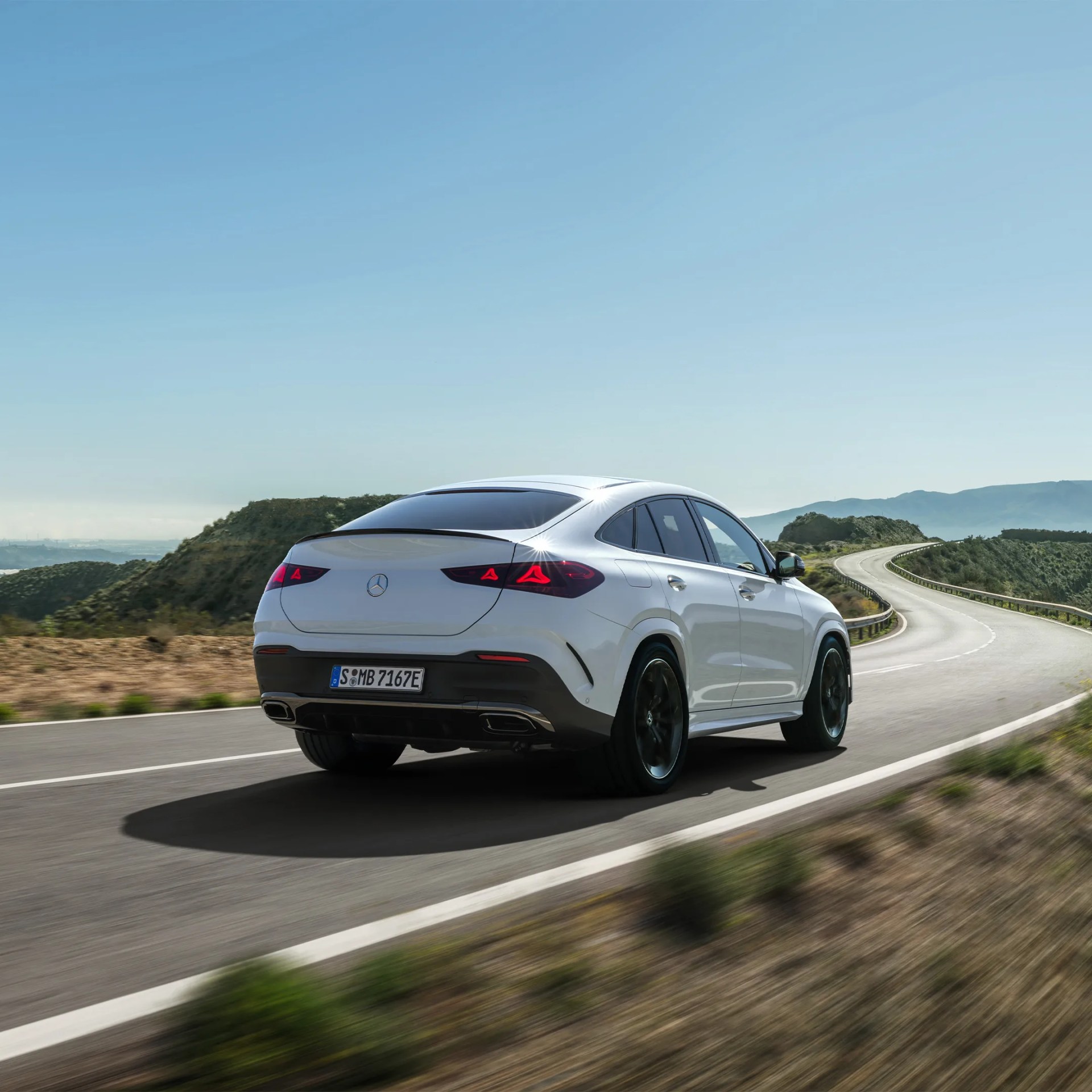 White Mercedes-Benz SUV coupe driving on a winding road with mountains in the background.