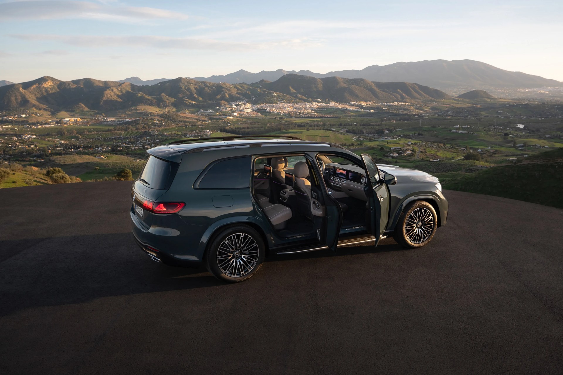Dark green Mercedes-Benz SUV with open front and rear passenger doors parked on a hilltop with mountains in the background.