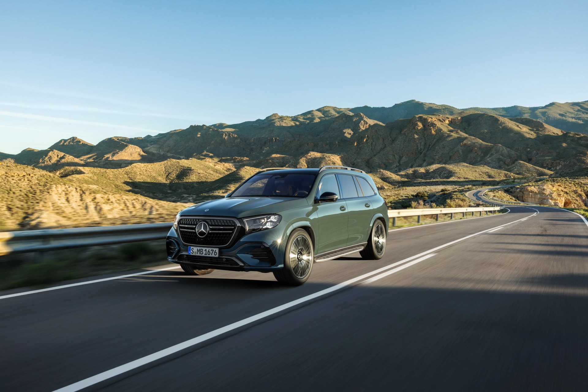Green Mercedes-Benz SUV driving on a winding mountain road under a clear blue sky.