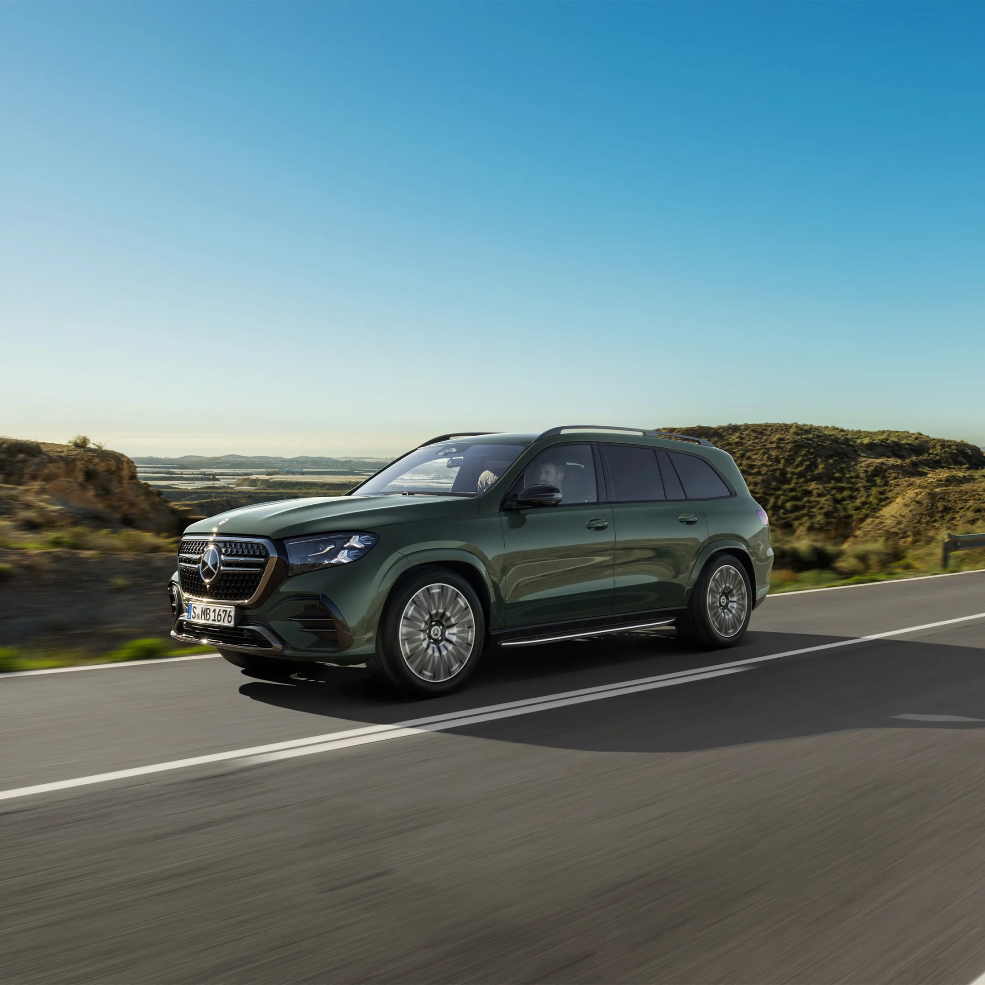 Dark green Mercedes-Benz SUV driving on a road with hills in the background under a clear blue sky.
