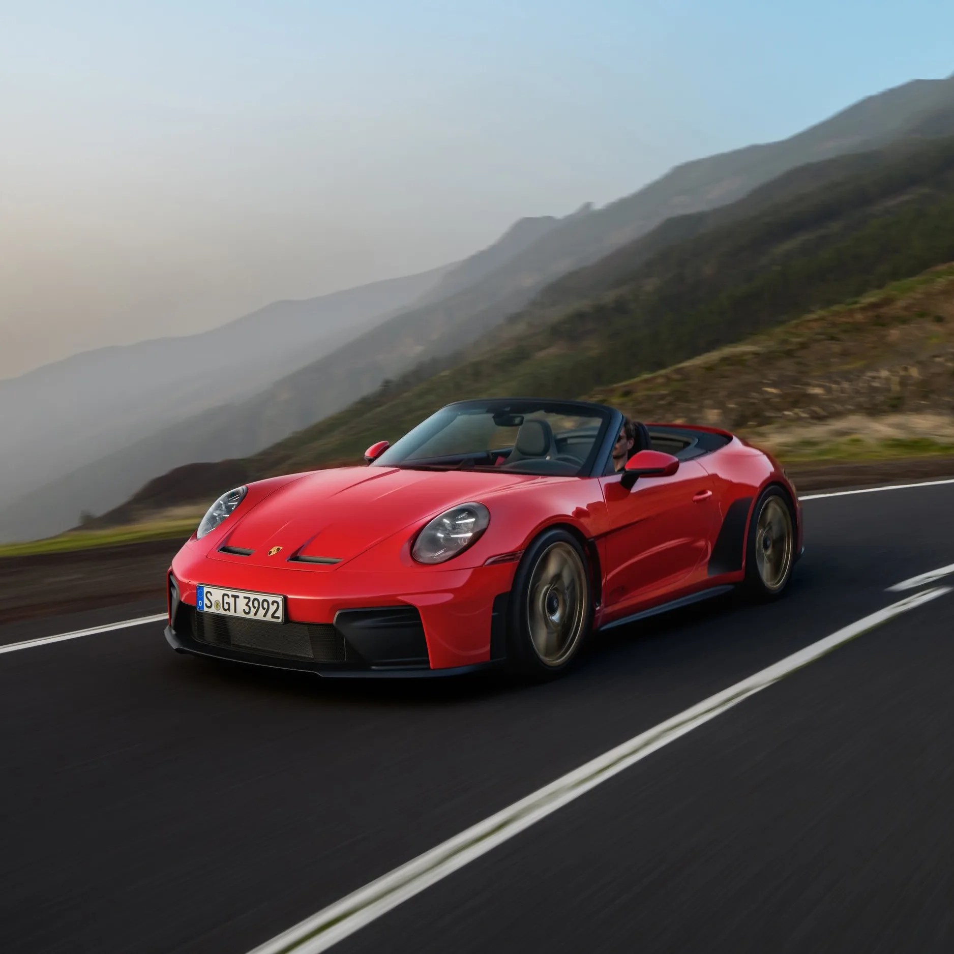 Red Porsche convertible sports car driving on a mountain road with a clear sky background.