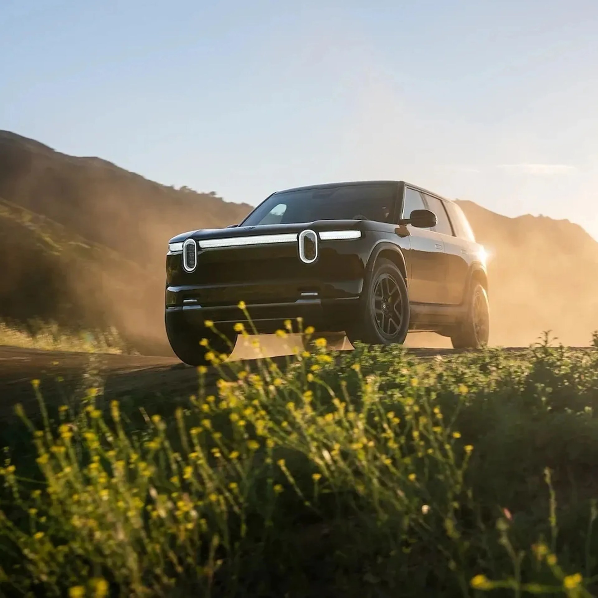 Black electric SUV driving on a dusty dirt road with hills in the background and yellow flowers in the foreground.