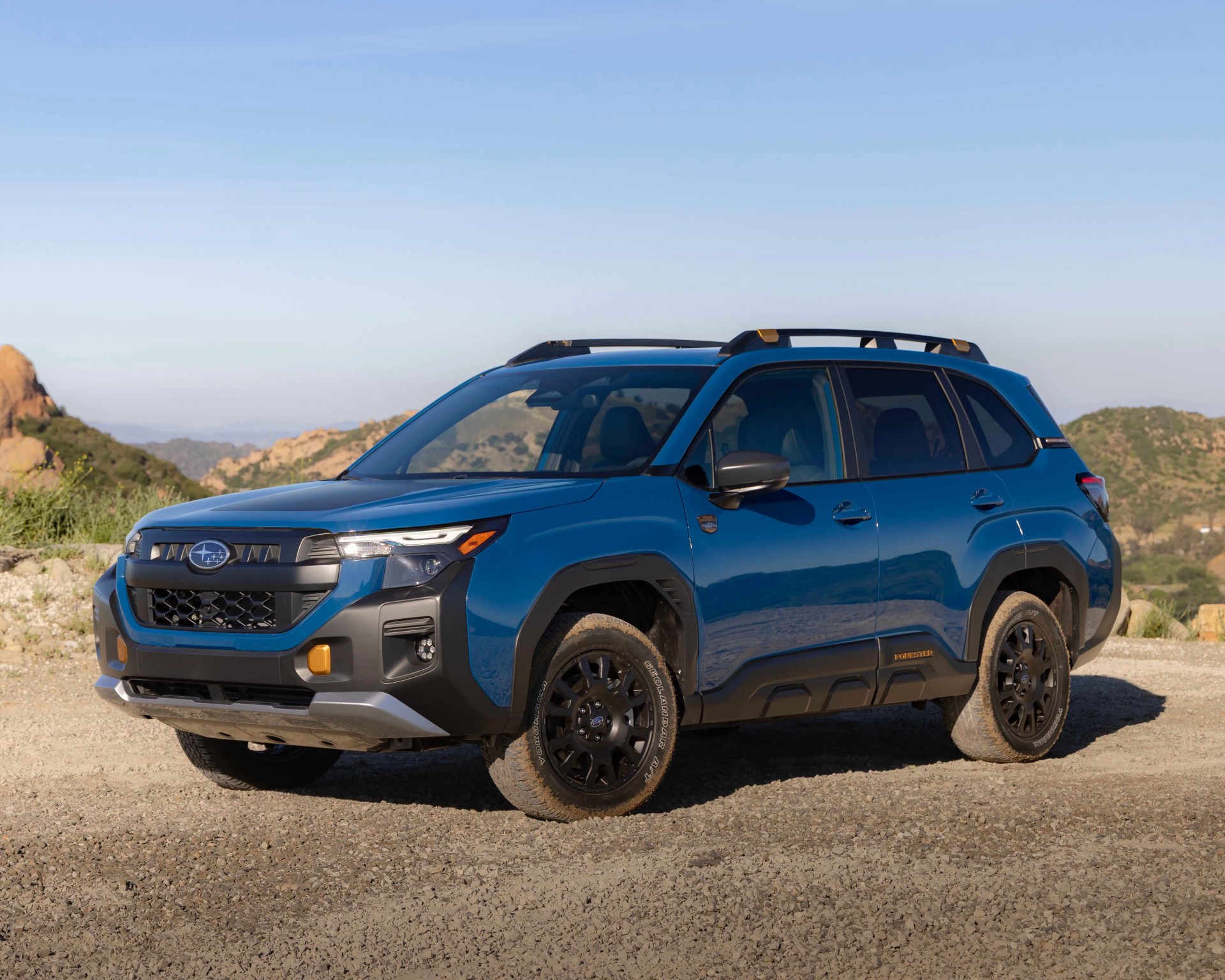 Blue Subaru Forester SUV with black and orange trim parked on a dirt road with hills in the background.