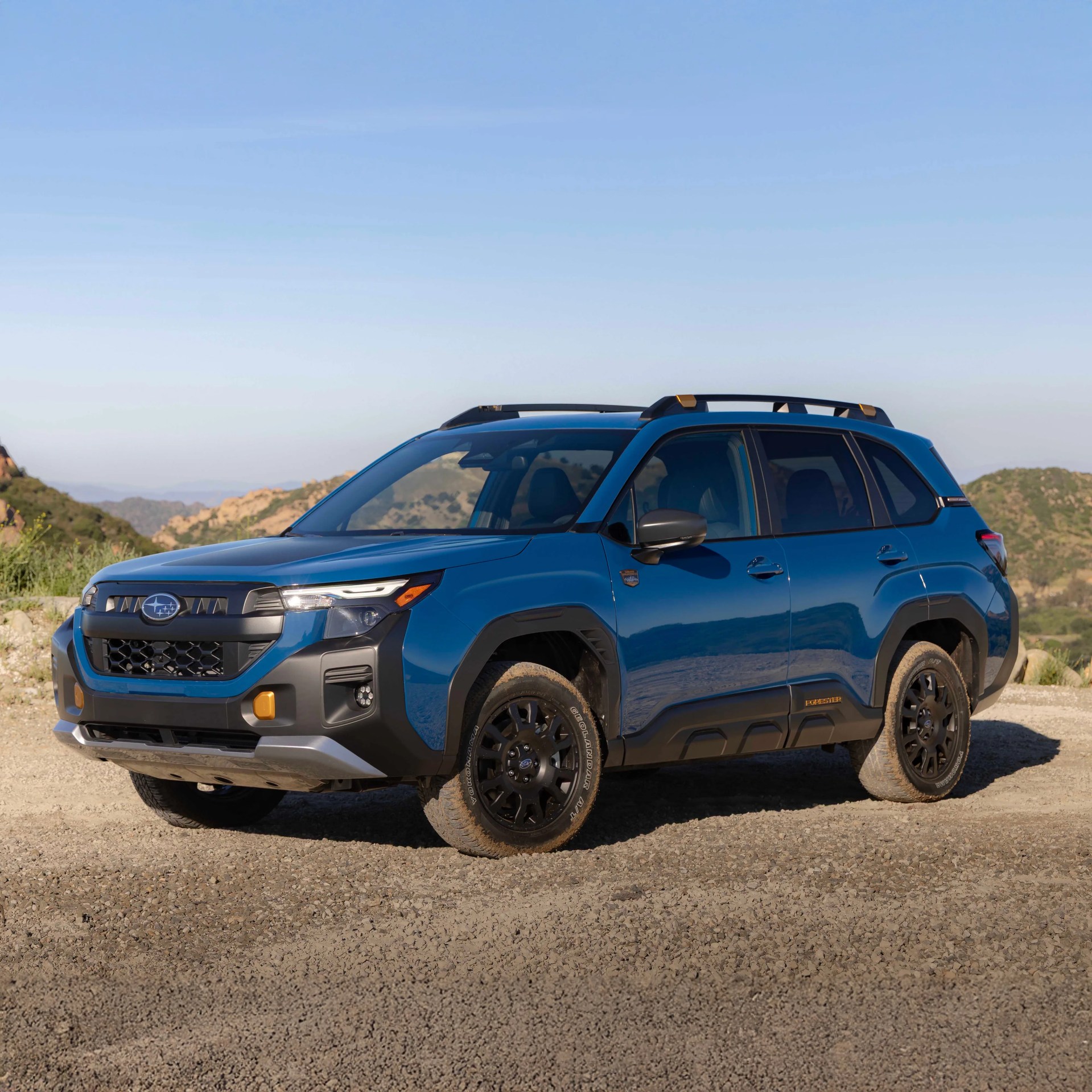 Blue Subaru SUV with black trim and roof rails parked on a dirt road with hills in the background.
