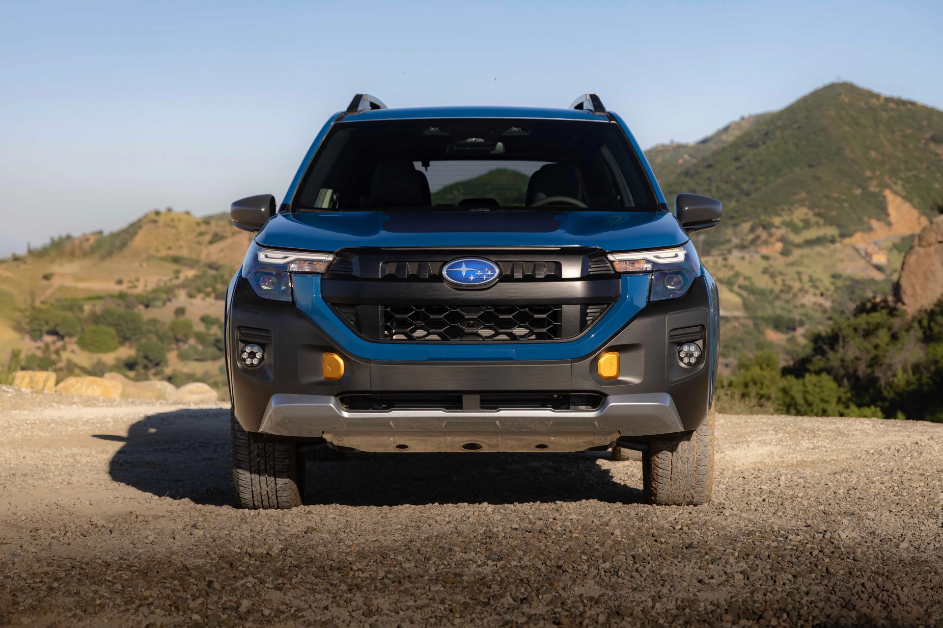 Front view of a blue Subaru SUV with black and silver accents parked on a gravel road with hills in the background.