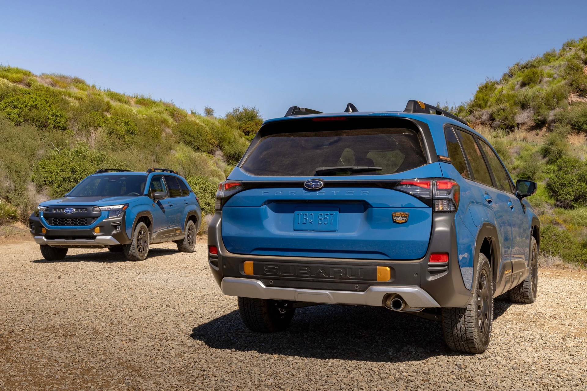 Two blue Subaru Forester SUVs parked on a gravel road with green hills in the background.