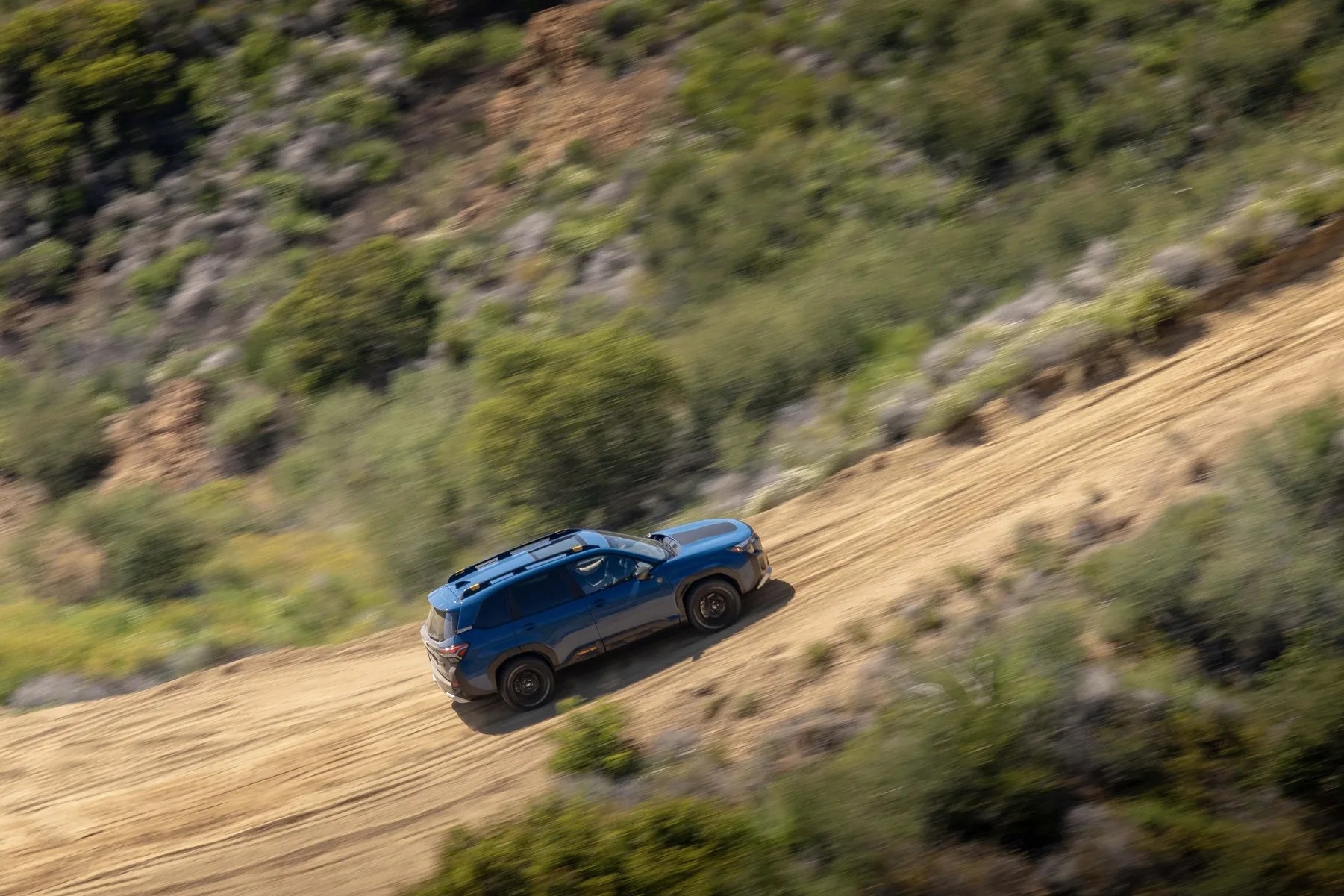 Blue SUV driving on a dirt road surrounded by green shrubs and rocky terrain.