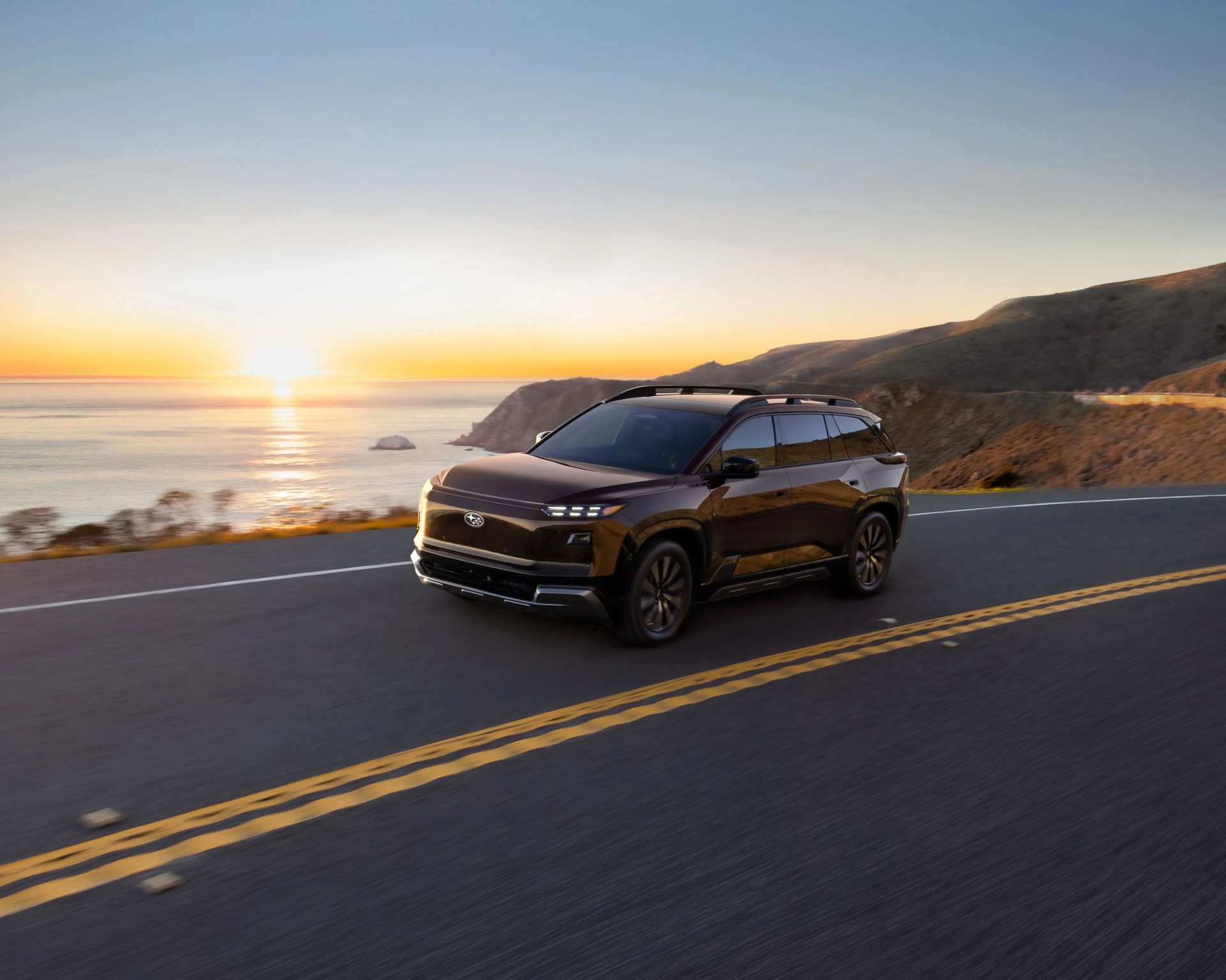 Dark brown Subaru SUV driving on coastal road at sunset with ocean and hills in background.