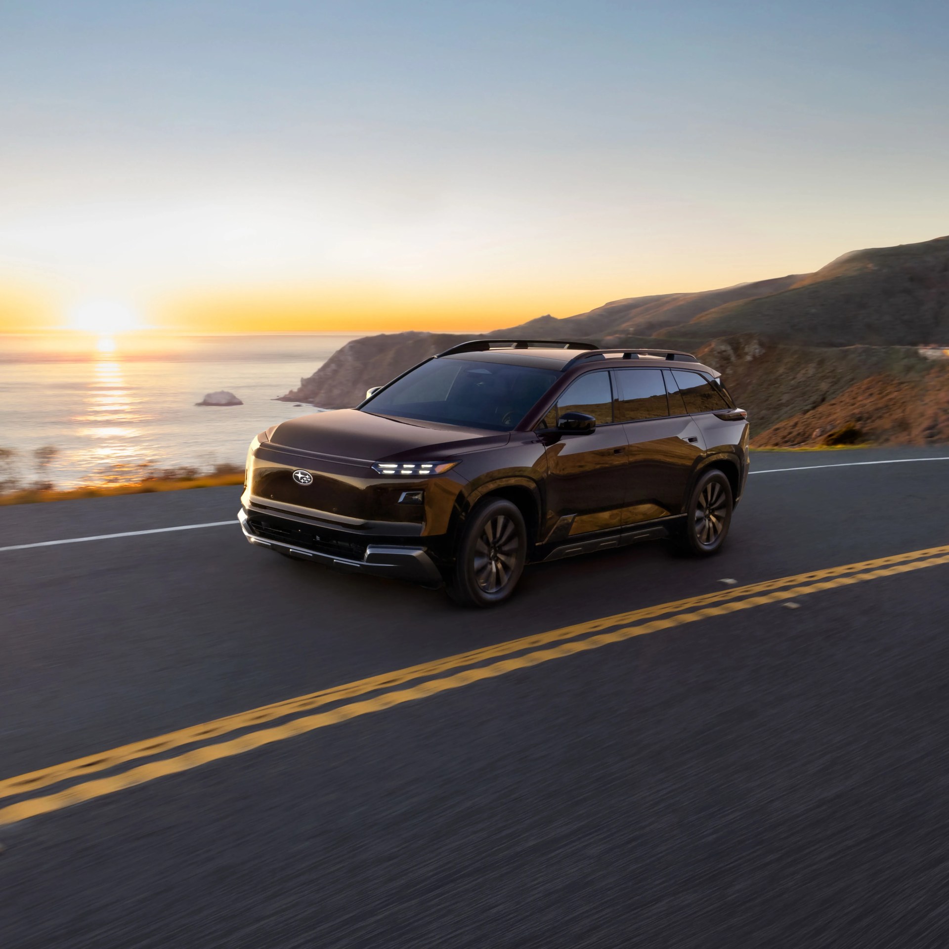Black Subaru SUV driving on coastal road at sunset with hills and ocean in background.