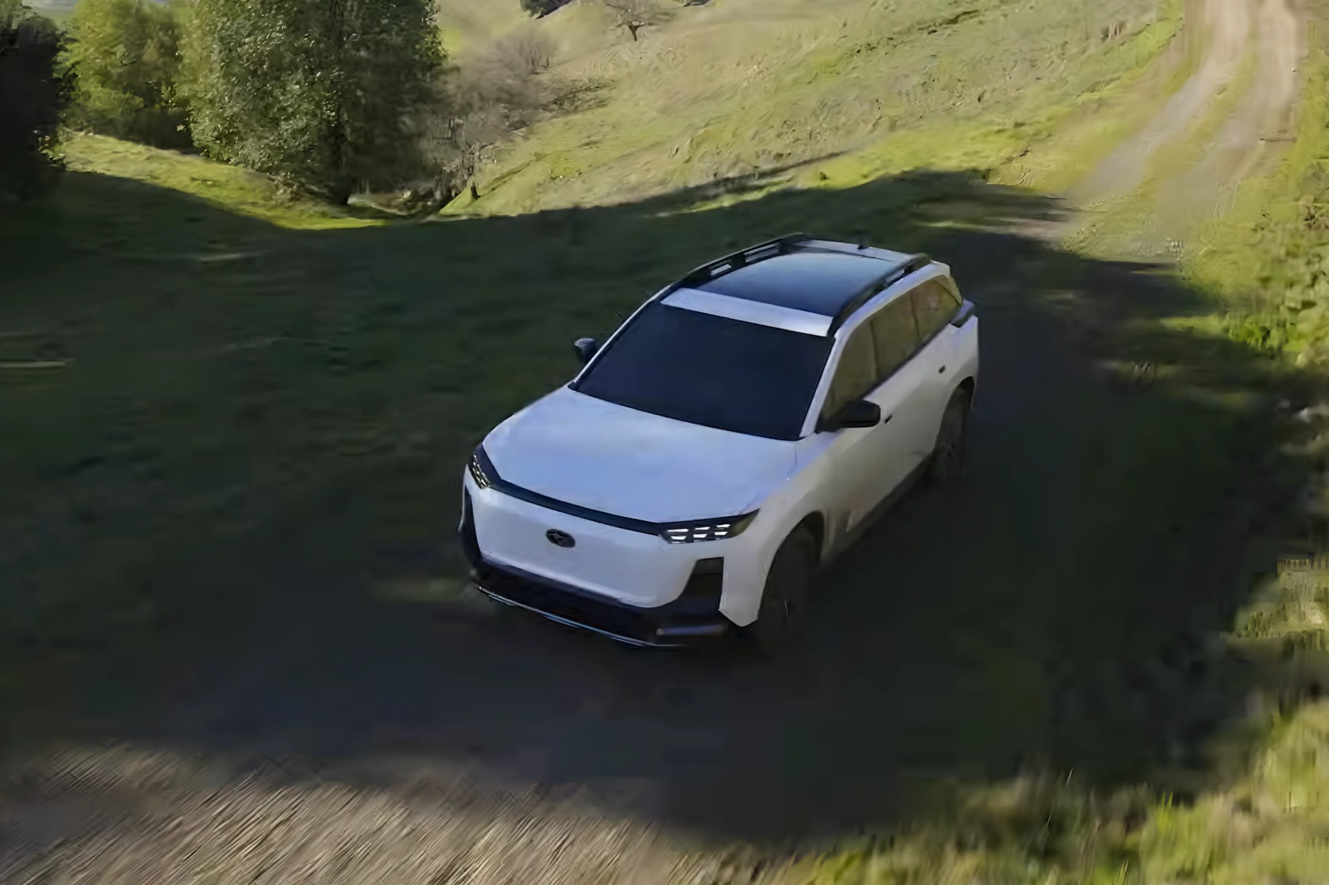 White electric SUV with black roof and tinted windows driving on a dirt road in a green hilly area.