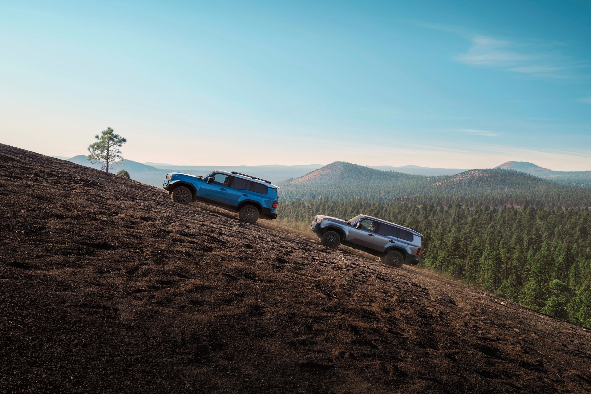 Two SUVs, one blue and one silver, driving uphill on a steep dirt slope with a forest and mountains in the background.