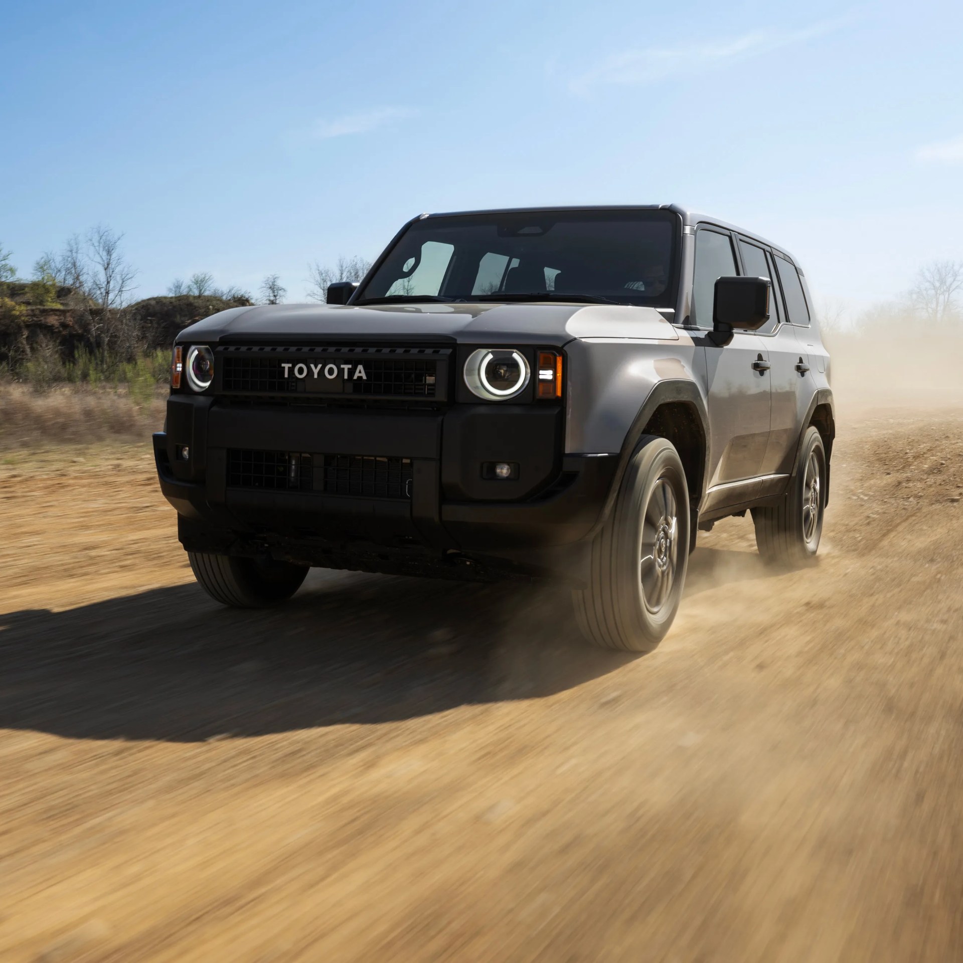 Silver Toyota SUV driving on a dusty dirt road with a clear blue sky background.