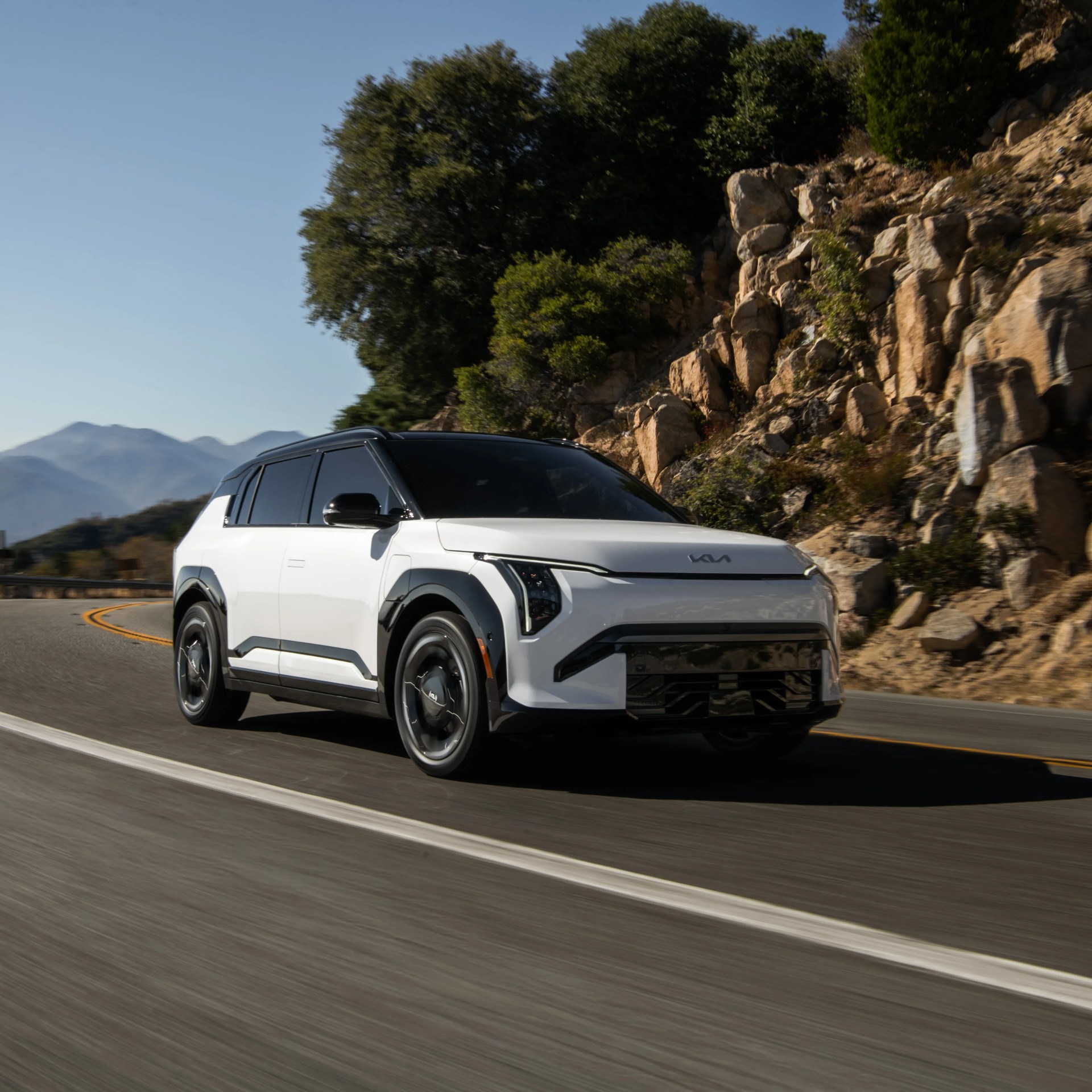 White Kia electric SUV with black roof and accents driving on a mountain road with rocky hillside and trees.