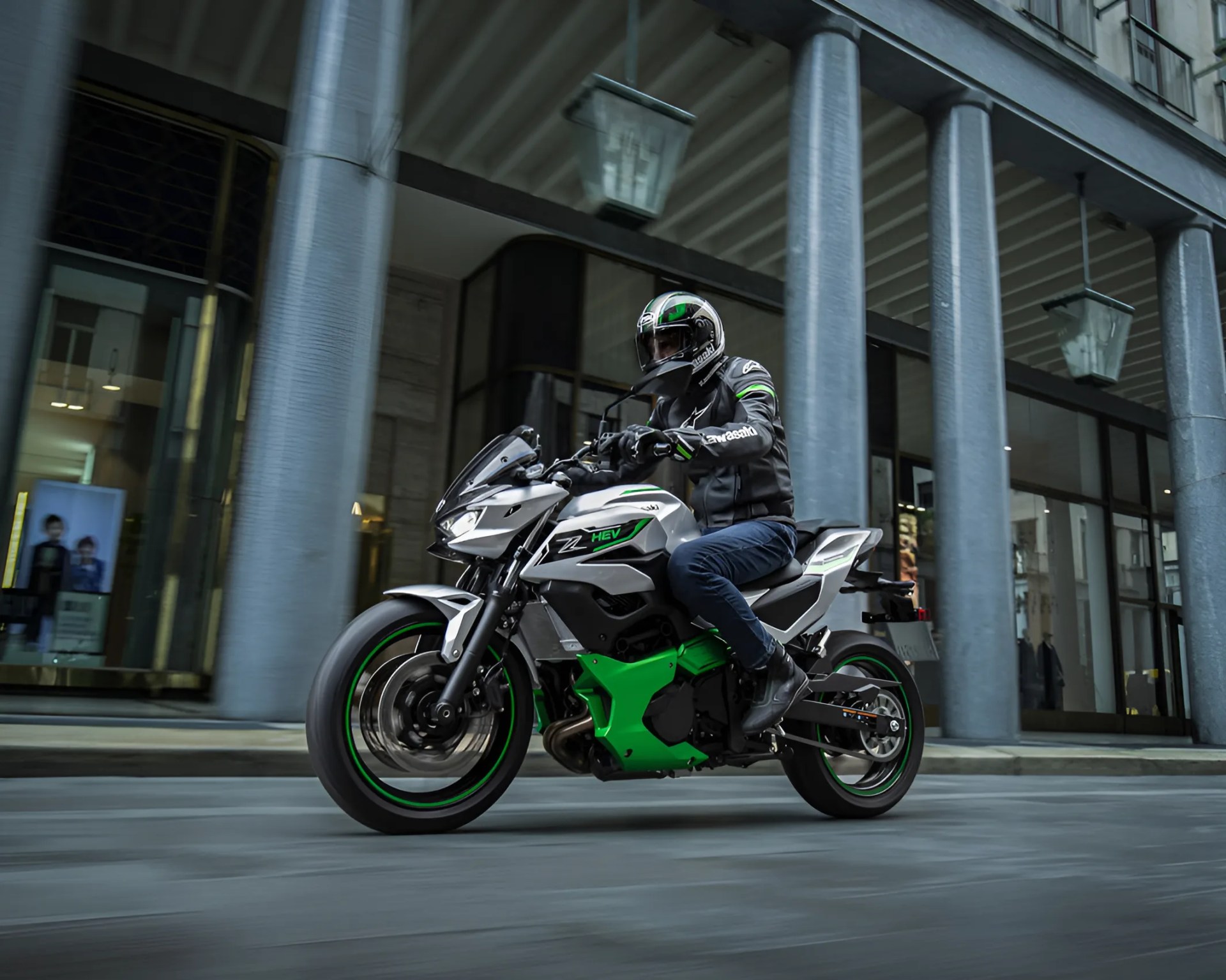 Person riding a silver and green sport motorcycle on a city street with tall gray columns in the background.
