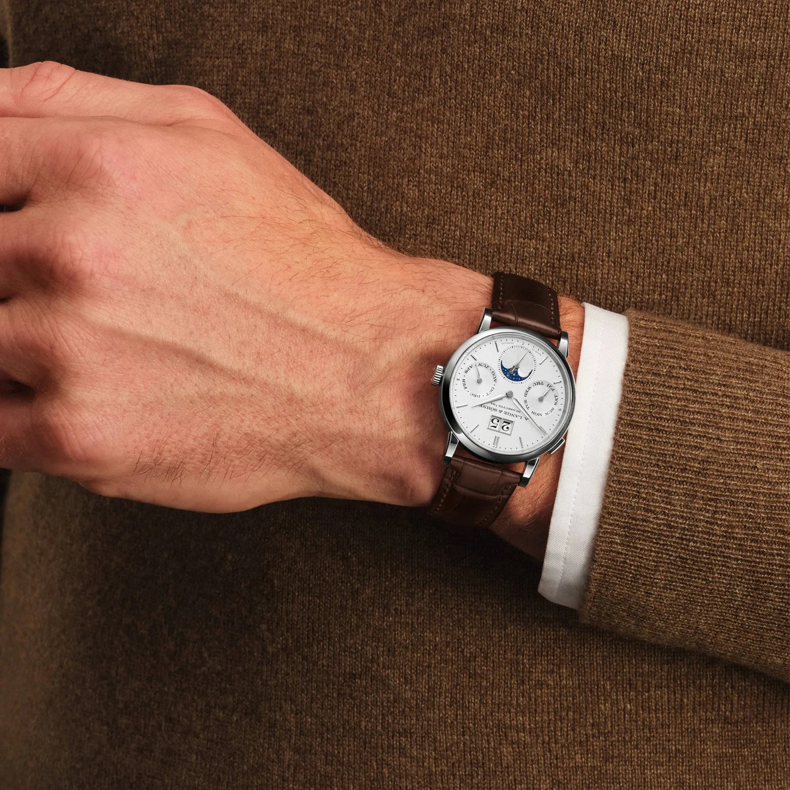 Silver wristwatch with white dial, moon phase, date display, and brown leather strap worn on a wrist over a brown sweater.