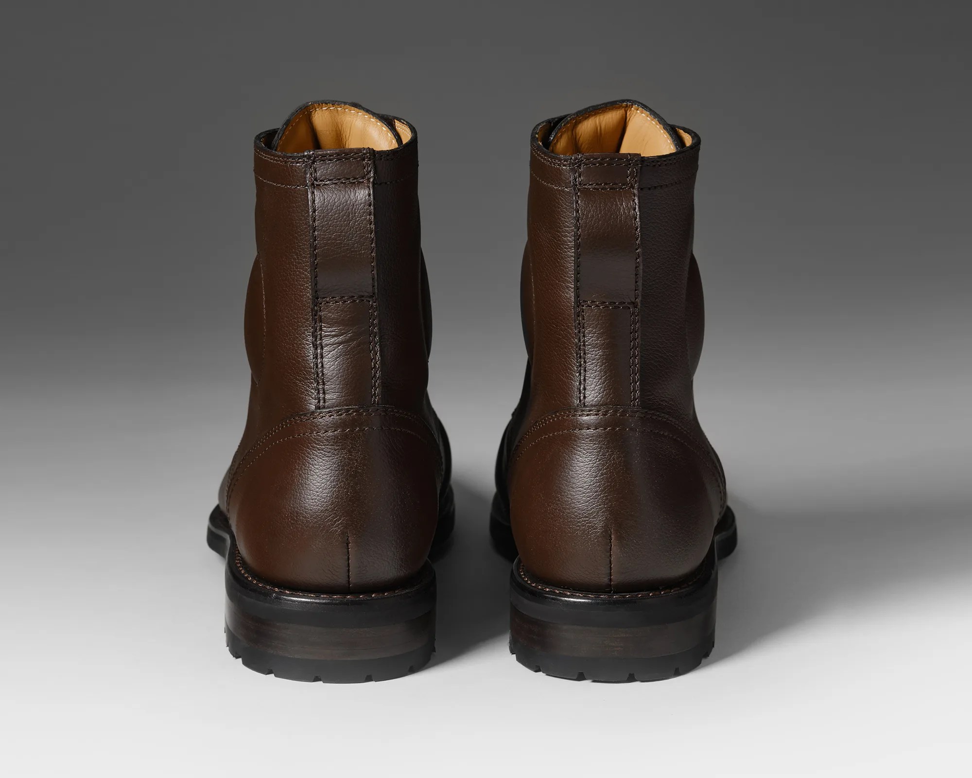 Pair of dark brown leather boots with black soles shown from the back on a gray background.