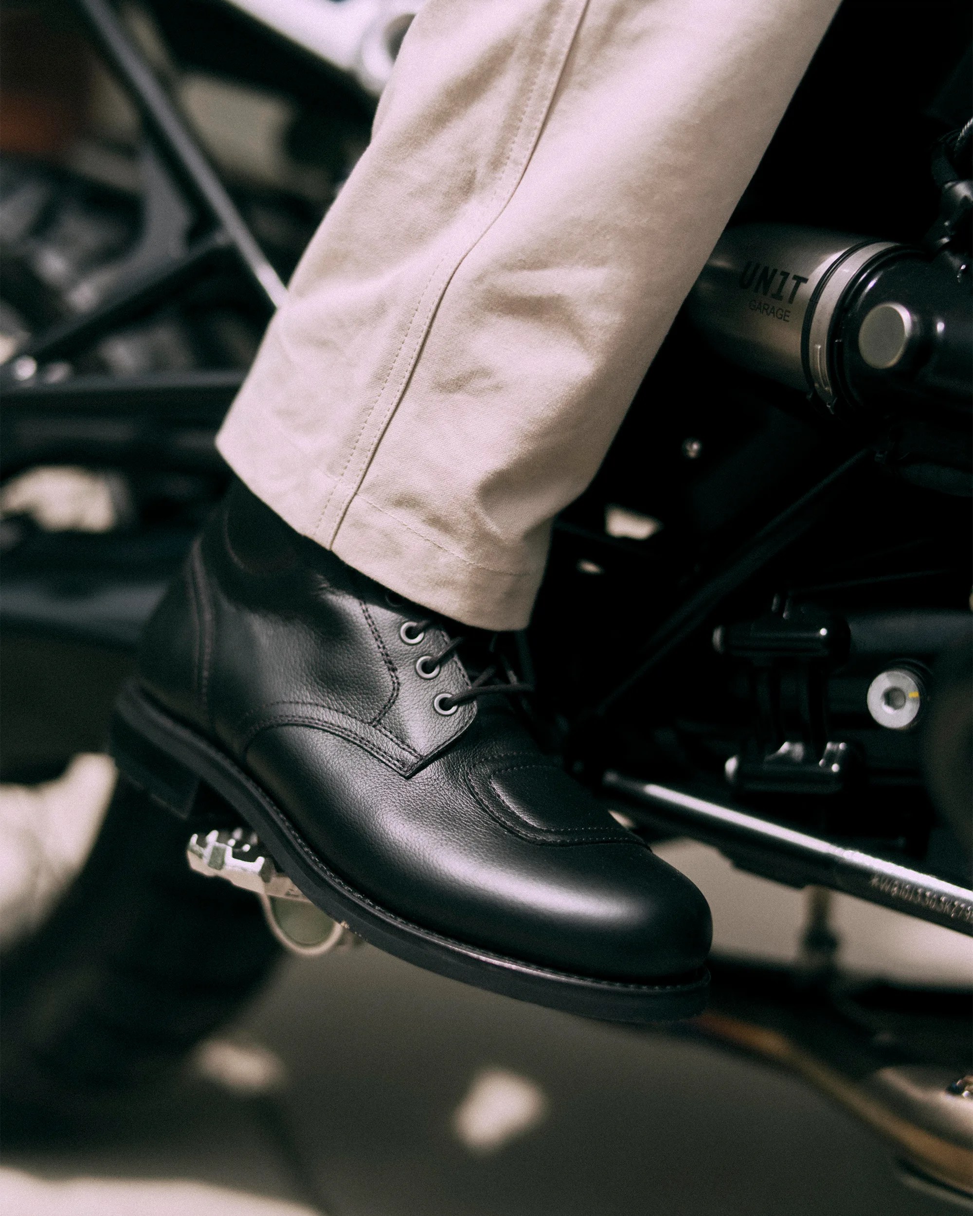 Black leather lace-up boot worn with beige pants resting on a motorcycle foot peg.