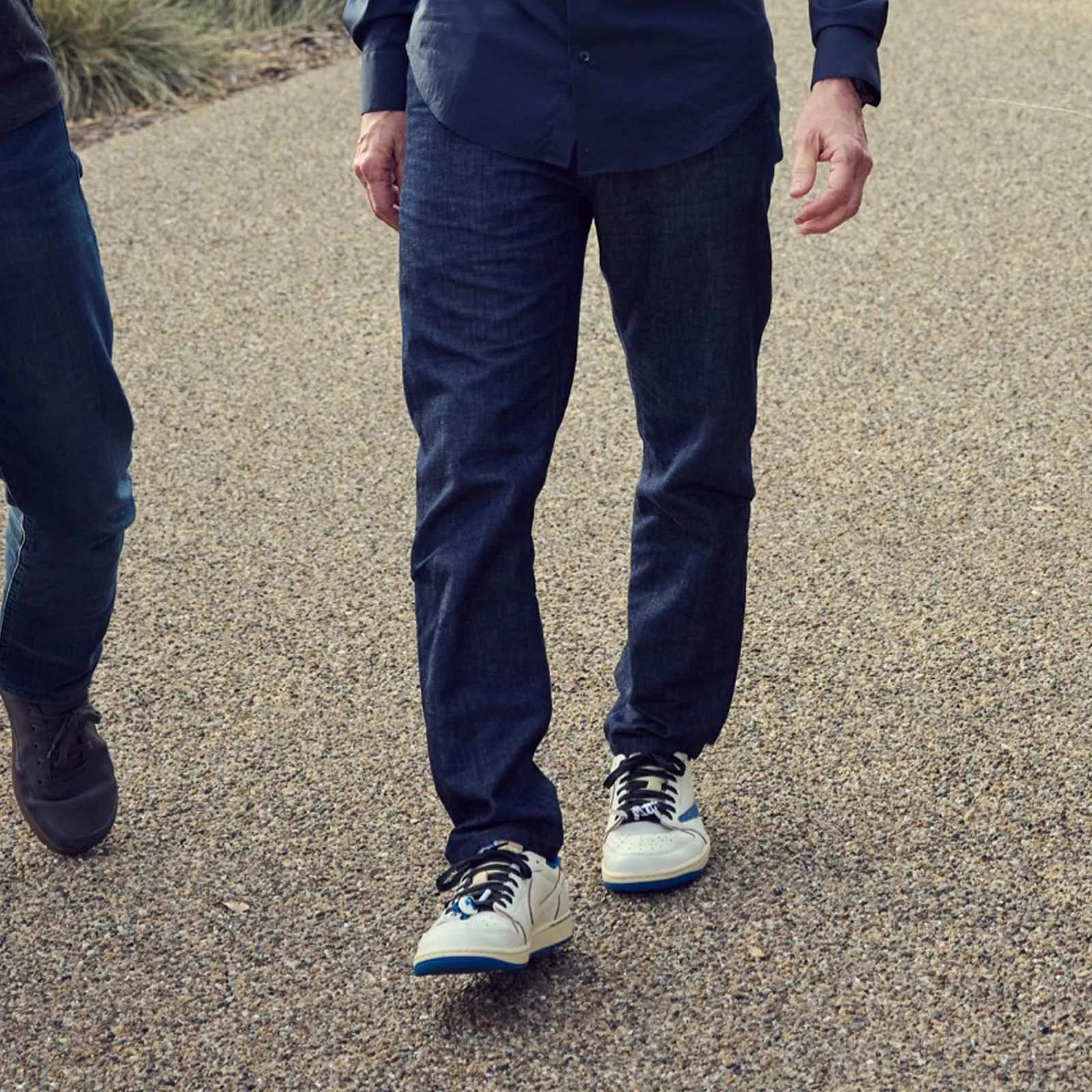 White sneakers with blue and black accents worn by a person in dark blue jeans walking on a gravel path.