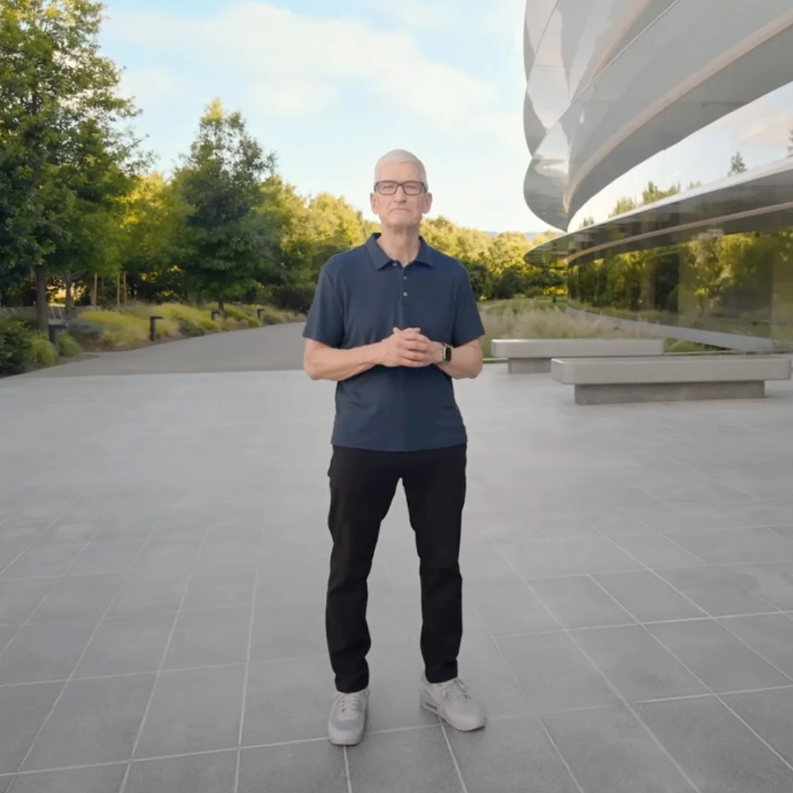 Person wearing a navy polo shirt, black pants, and white sneakers standing on a tiled outdoor area near a modern building.