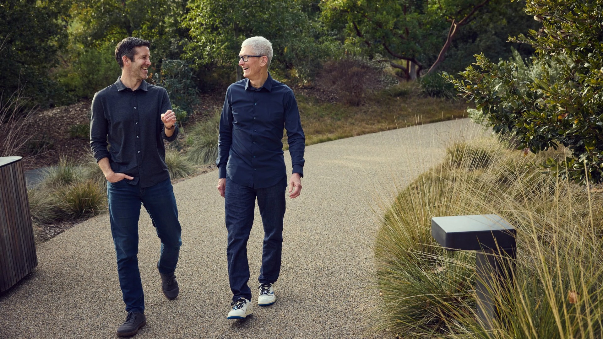 Two men walking and smiling on a gravel path surrounded by greenery and bushes.