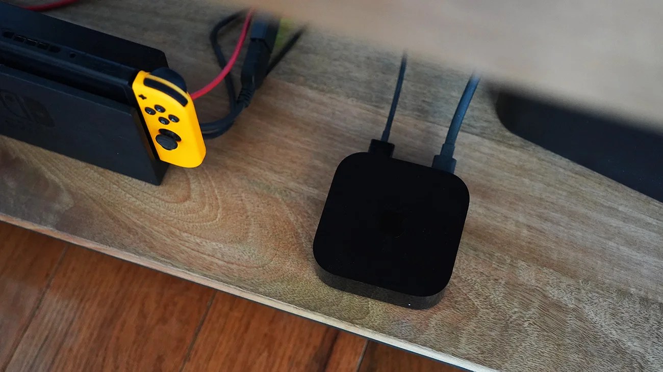 Black Apple TV device with two cables connected, placed on a wooden surface next to a Nintendo Switch dock with a yellow Joy-Con.