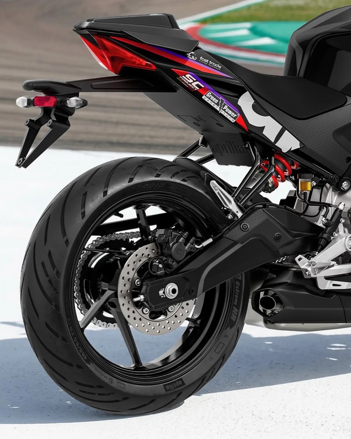 Rear section of a black Aprilia motorcycle showing the rear tire, disc brake, red suspension spring, and tail light with decals.