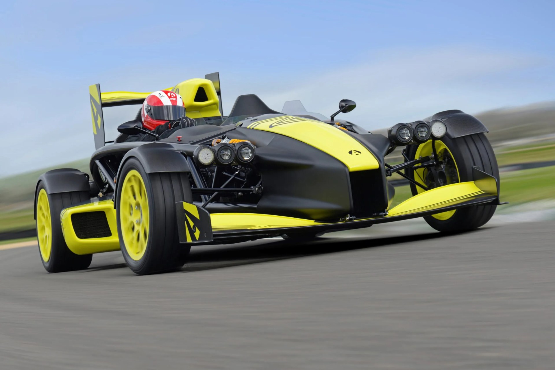 Black and yellow Ariel Atom open-wheel race car with a driver wearing a red and white helmet on a racetrack.