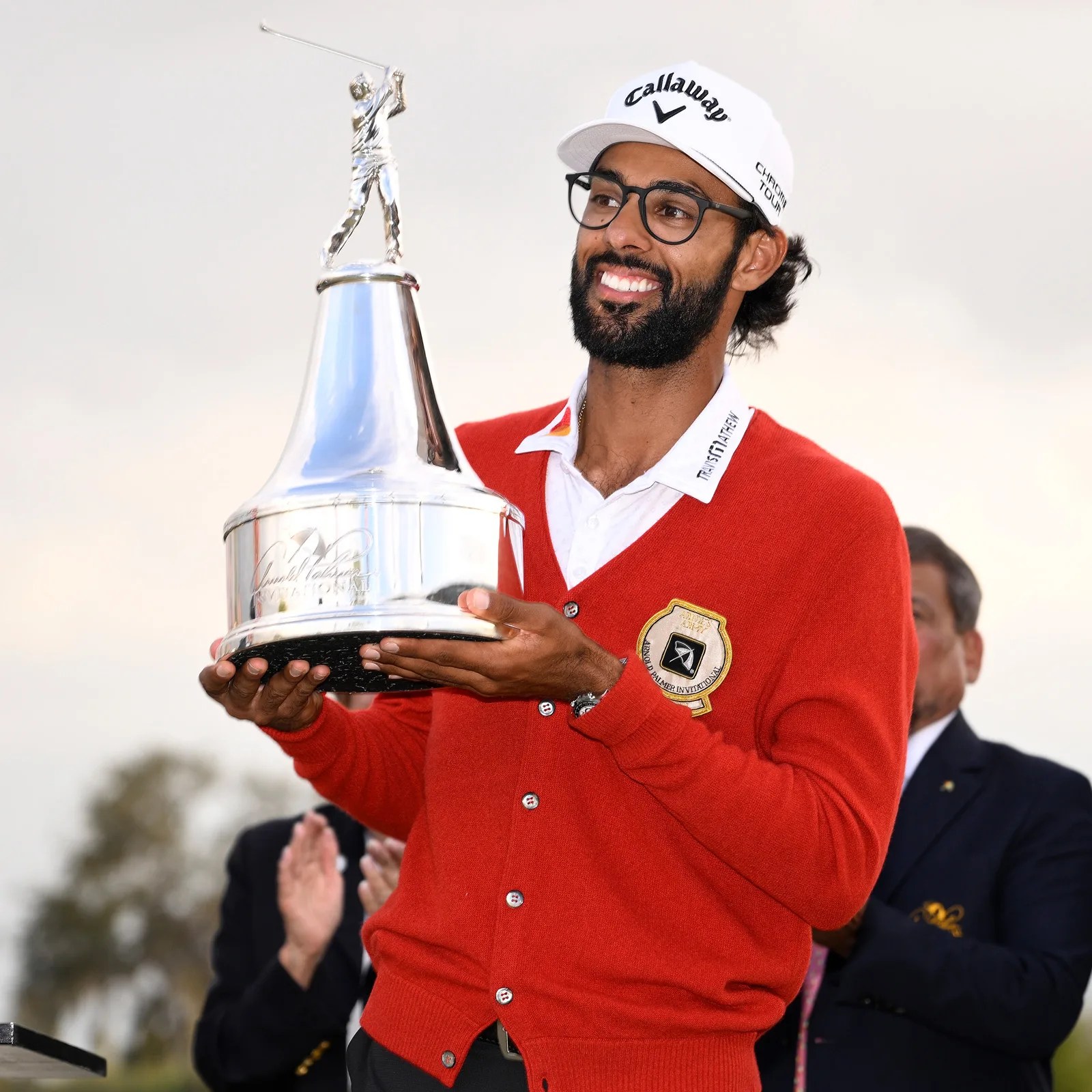 Man in a red cardigan and white Callaway cap holding a large silver golf trophy with a golfer figure on top.