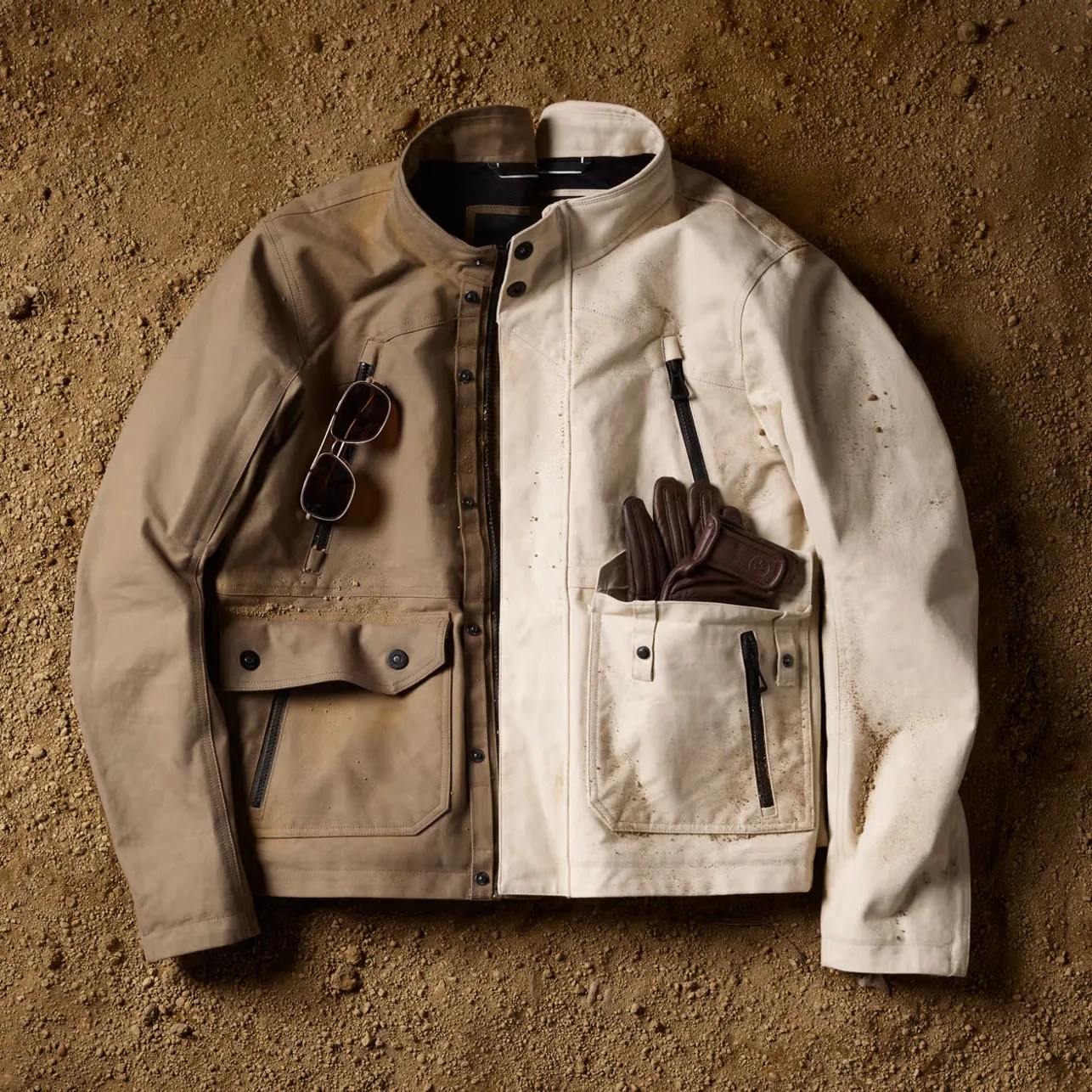Two-tone brown and off-white canvas jacket with front pockets, brown leather gloves in pocket, and sunglasses clipped on pocket, laid on dirt ground.