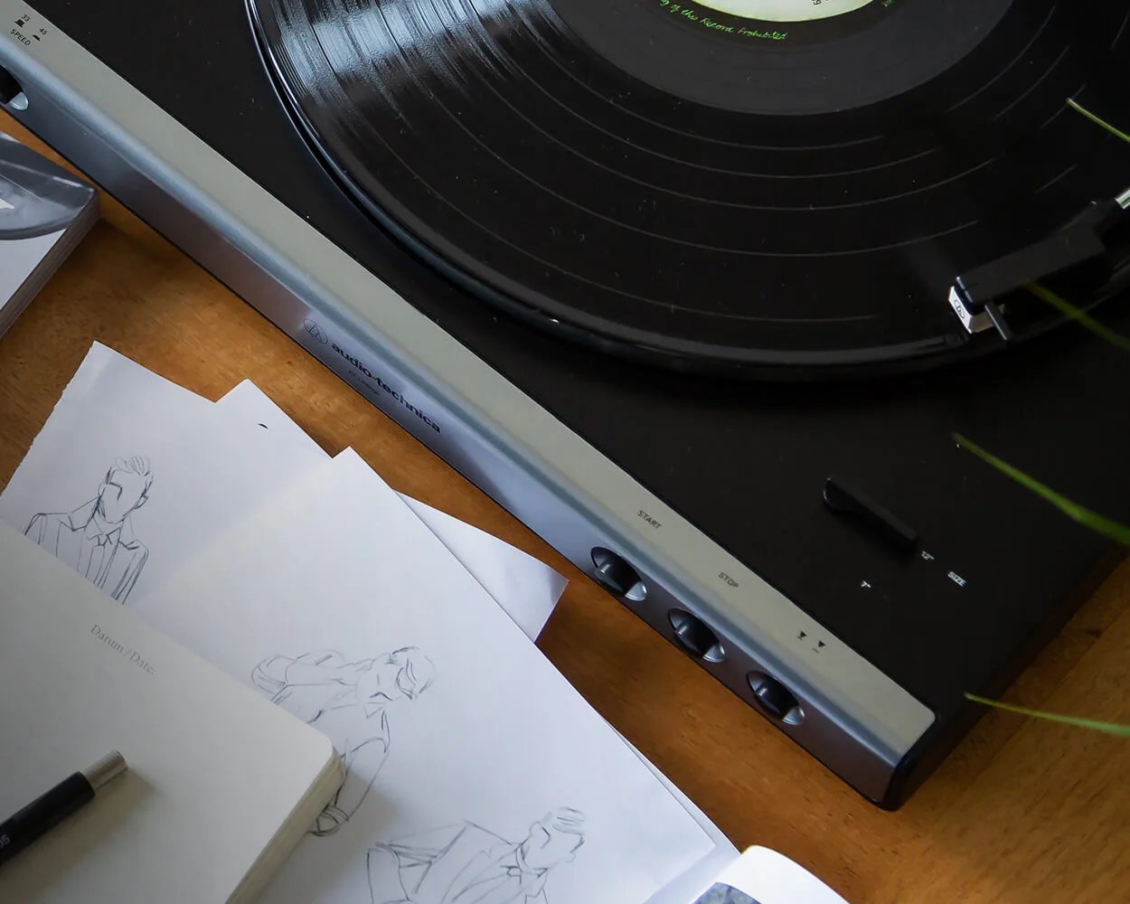 Black Audio-Technica turntable with a vinyl record on a wooden surface next to sketches of people on paper.