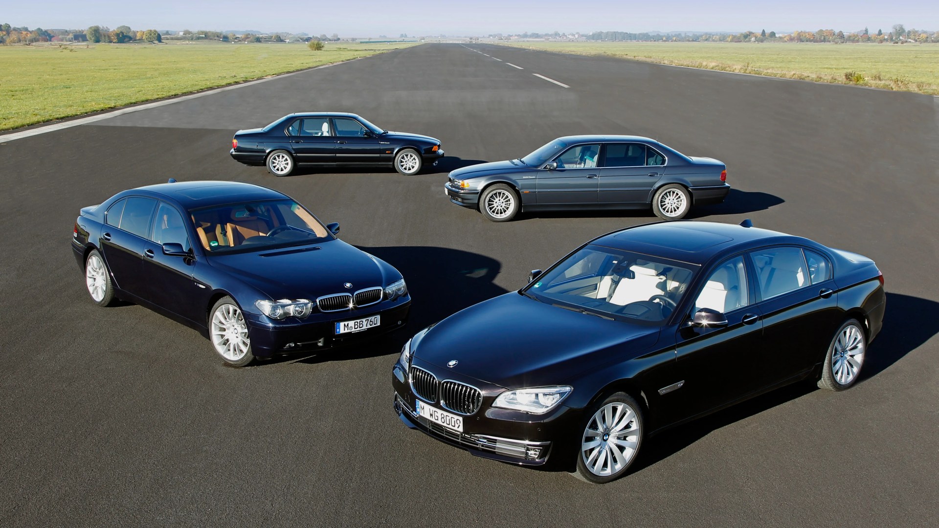 Four black BMW sedans parked on an open tarmac runway with green fields in the background.