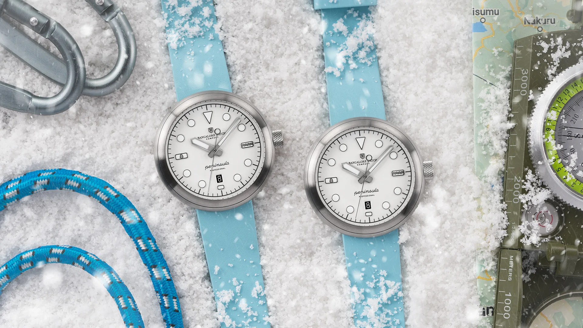 Two silver wristwatches with white dials and light blue straps on snow next to climbing gear and a compass.