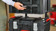 Hand lifting the top section of a black Bauer modular tool storage box with red and silver latches.
